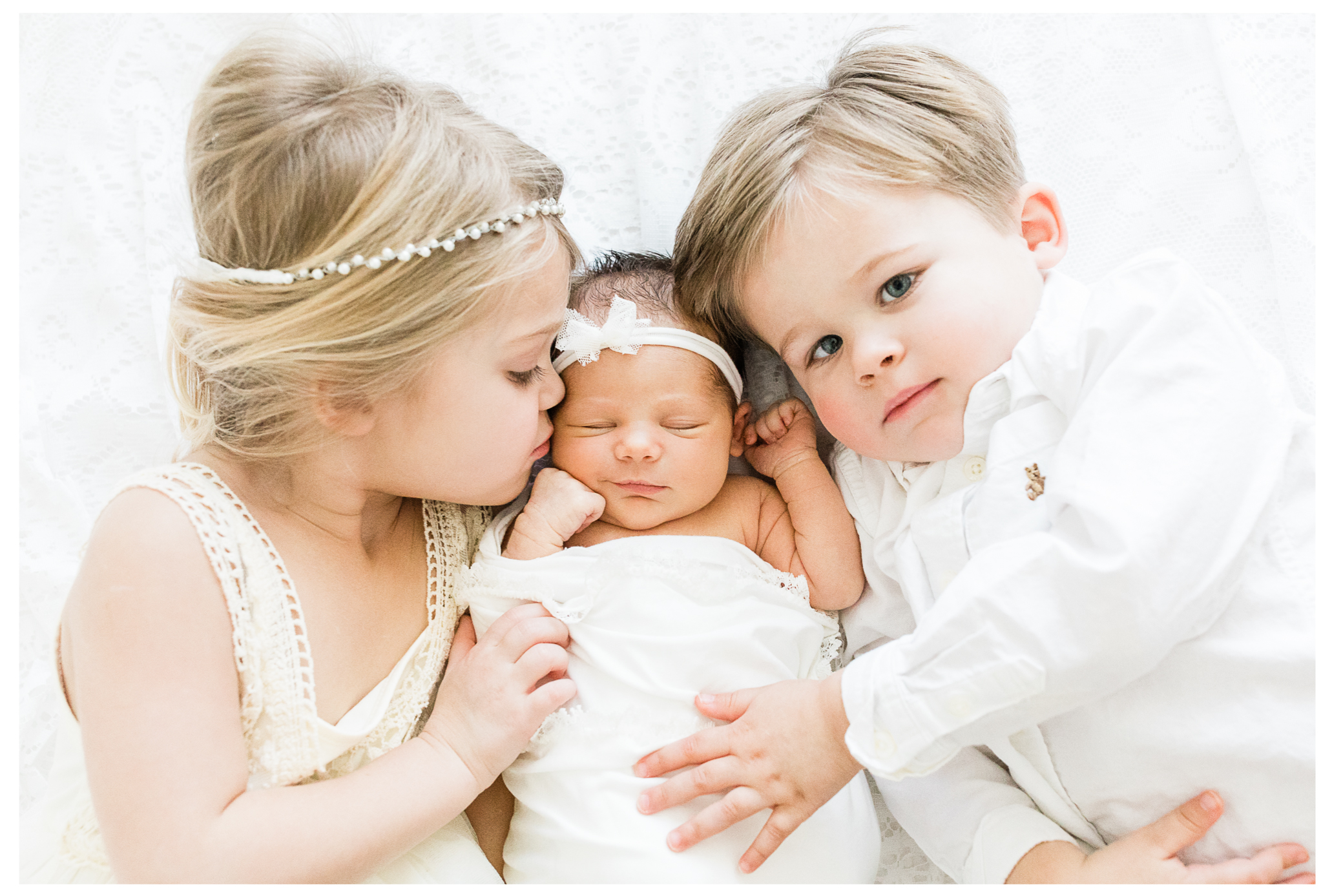 Two toddler siblings snuggling their newborn sister between them on a white bed