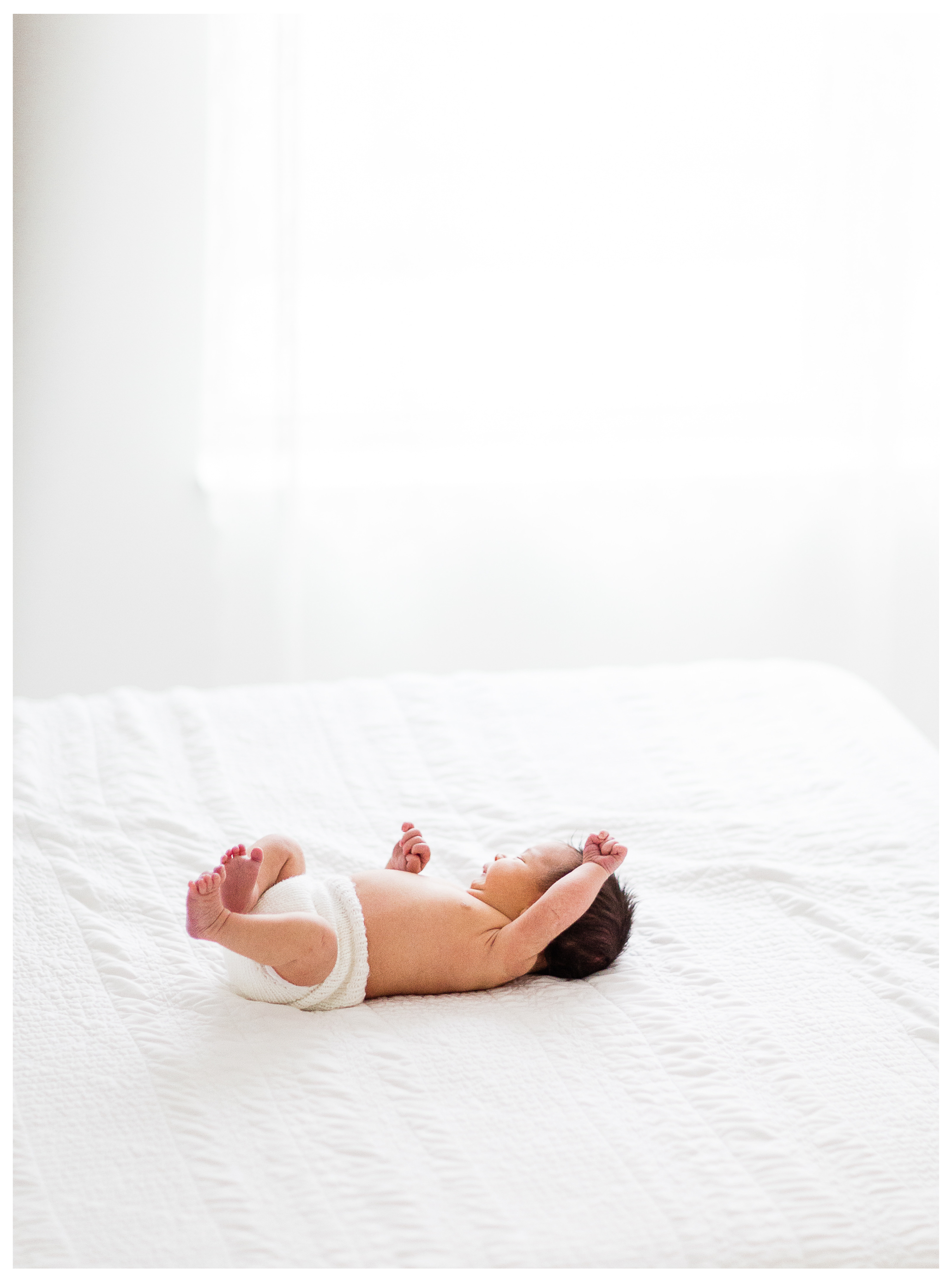 A newborn boy in a knit bloom stretching on a white studio bed in the window light