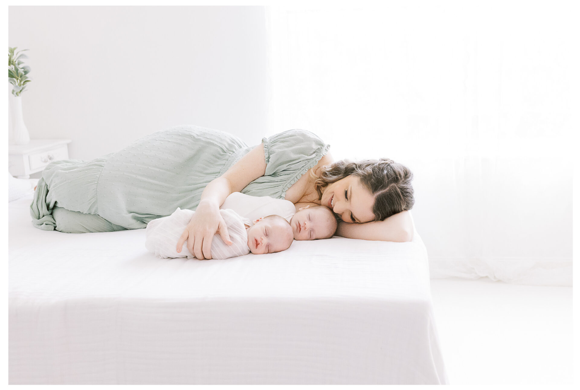 Twin baby boy newborn photography session | Mother backlit with dreamy light kissing her babies on a white bed