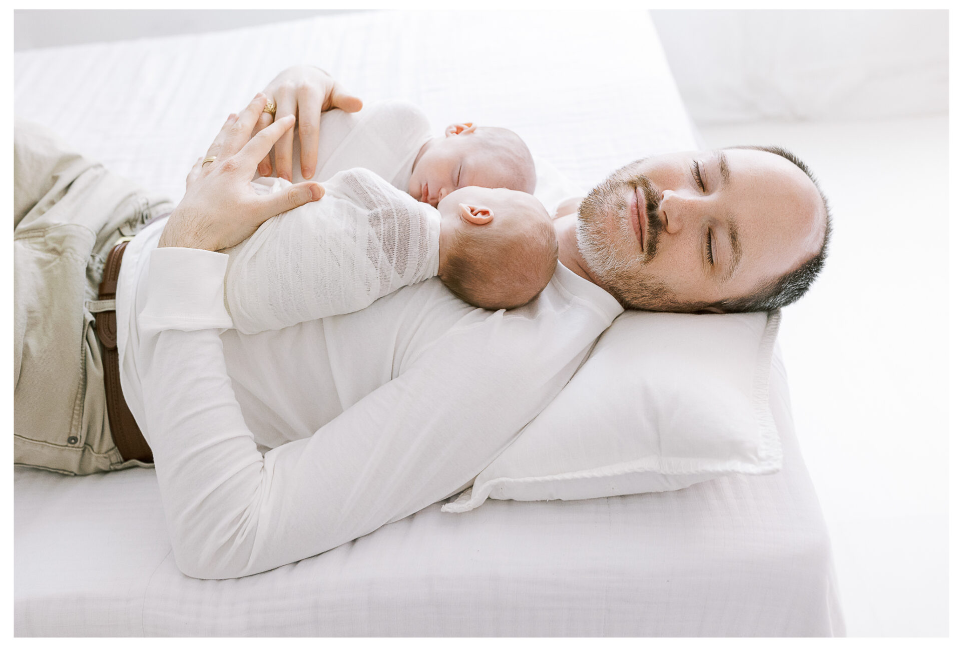 Dad snuggling his twin newborn baby boys on a white bed