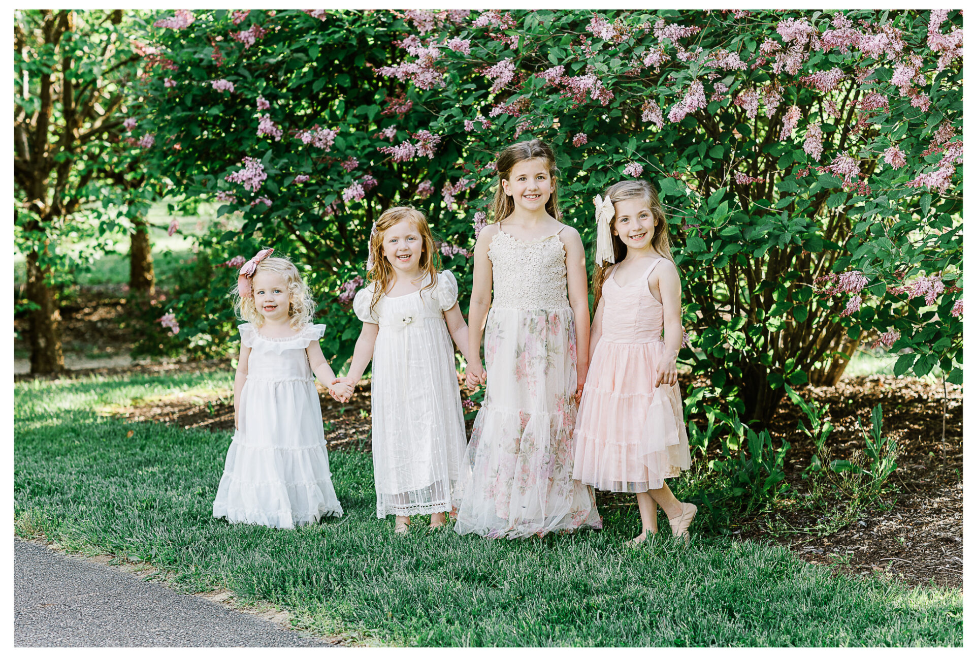 Four young sisters smiling together beside a pink blossoming bush in the springtime.