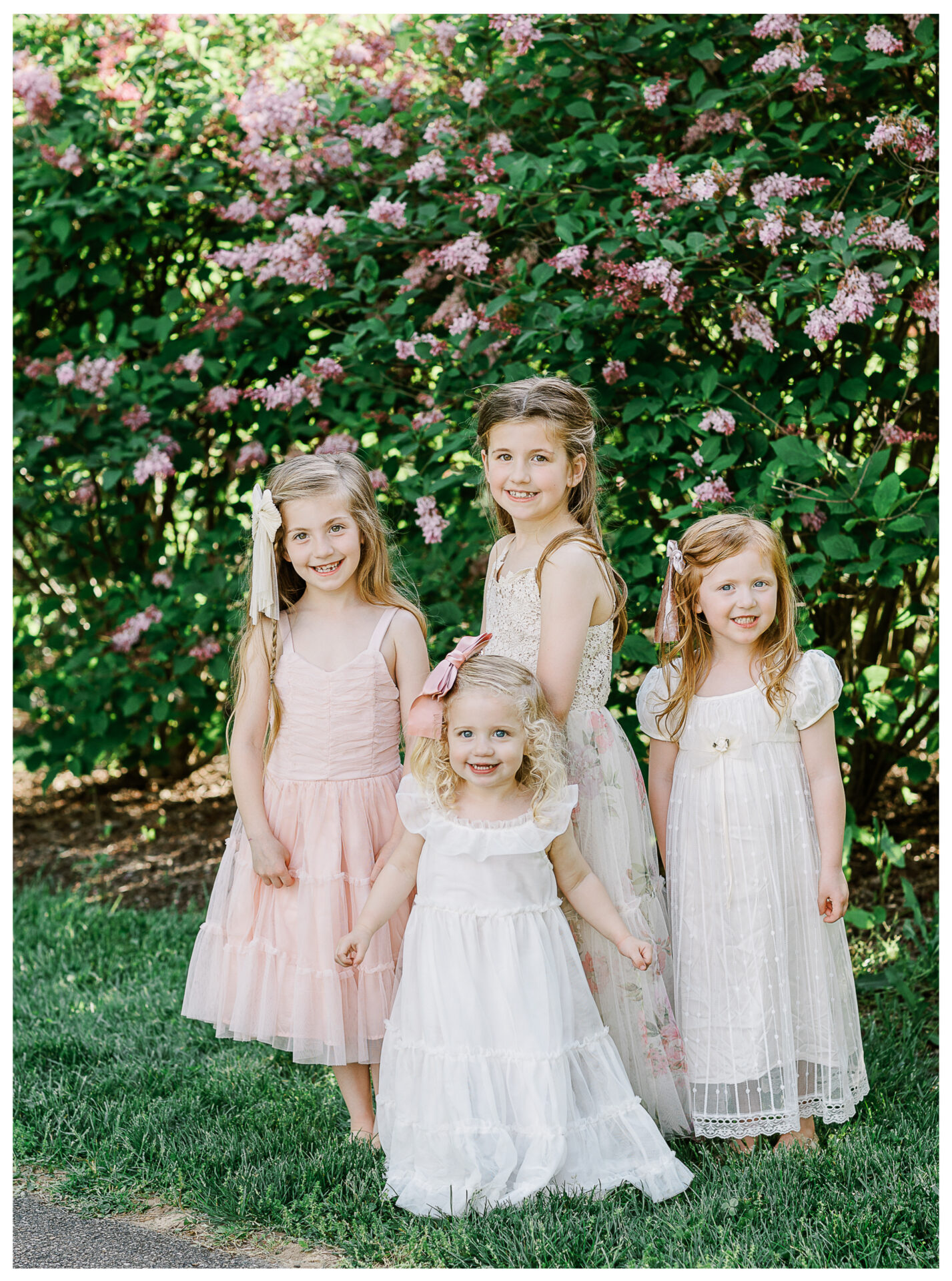 Four young sisters smiling together beside a pink blossoming bush in the springtime.