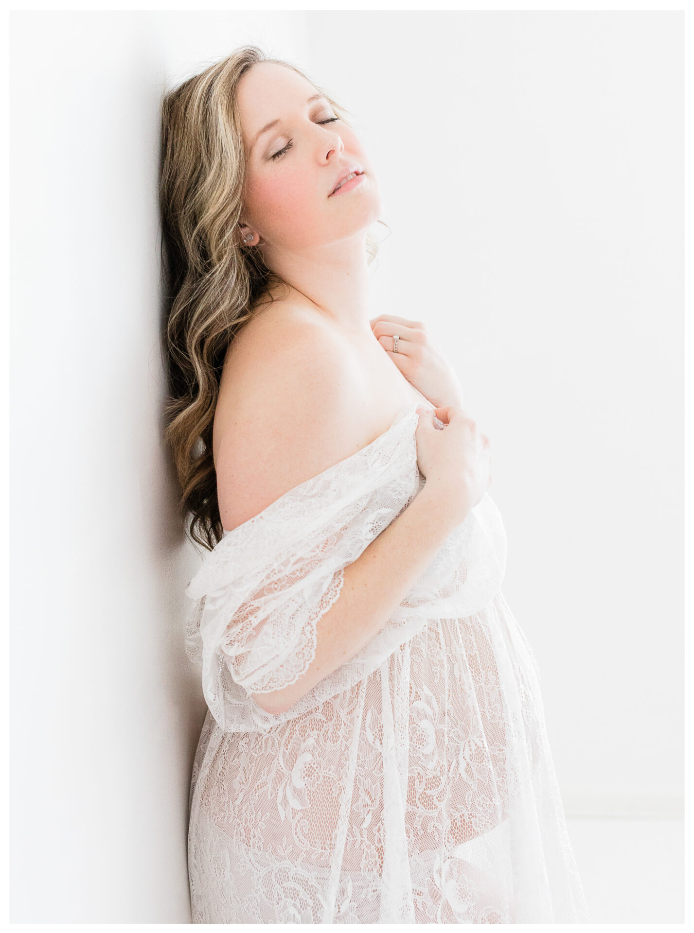 An expecting mother wearing lace and leaning gently against a white wall
