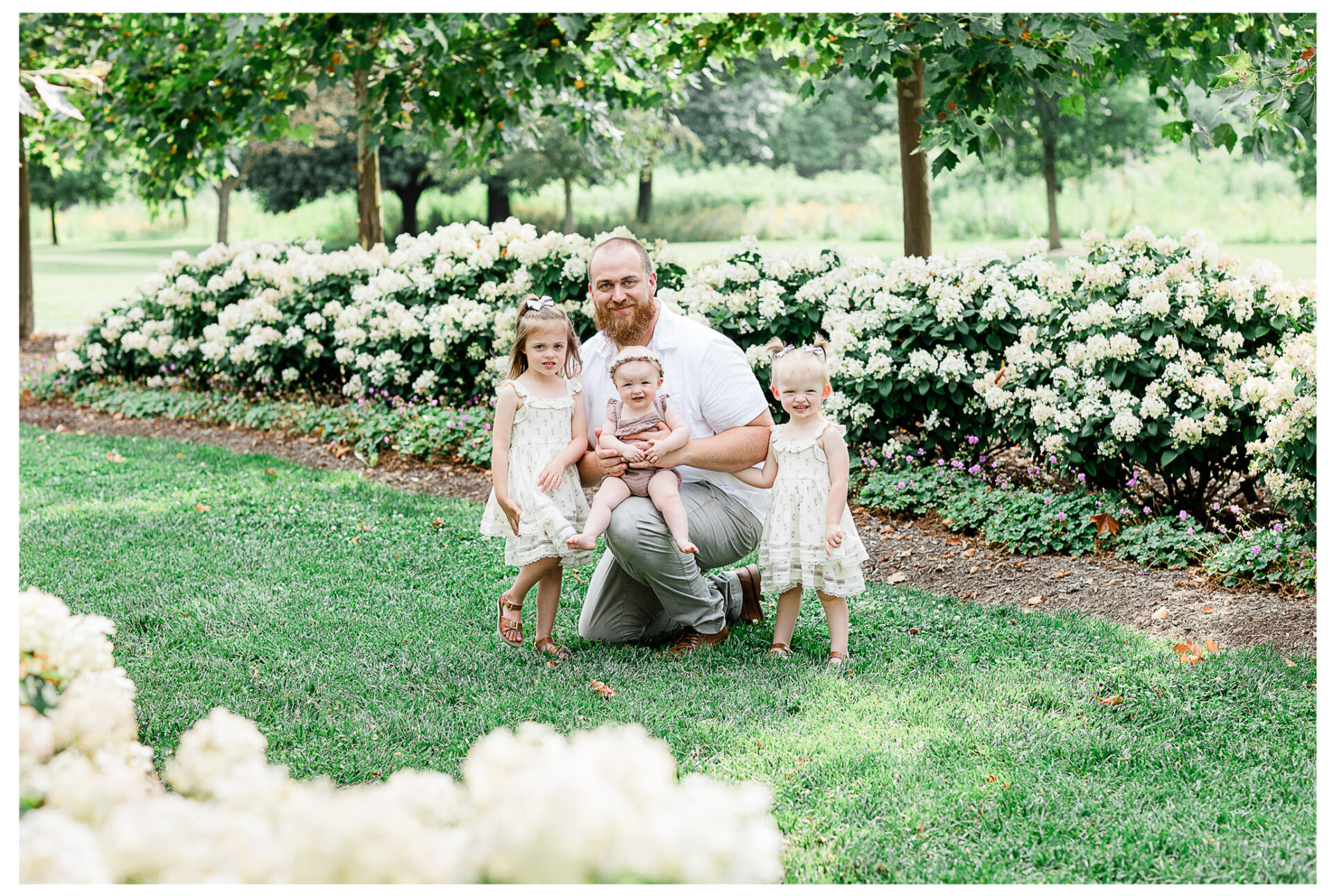 A father snuggling his three young daughters outside near flowering bushes.