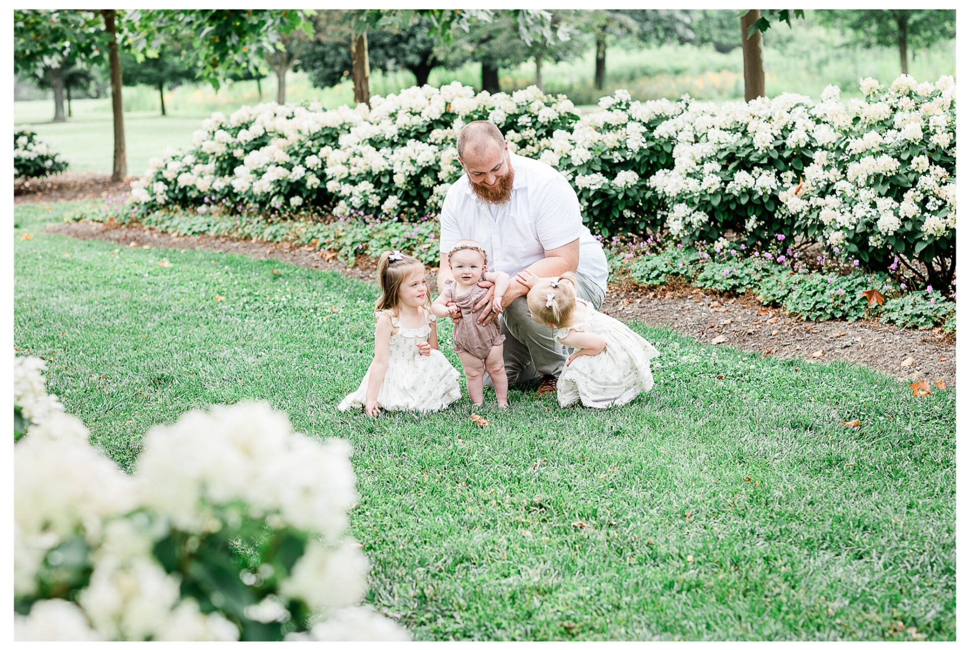 A father snuggling his three young daughters outside near flowering bushes.