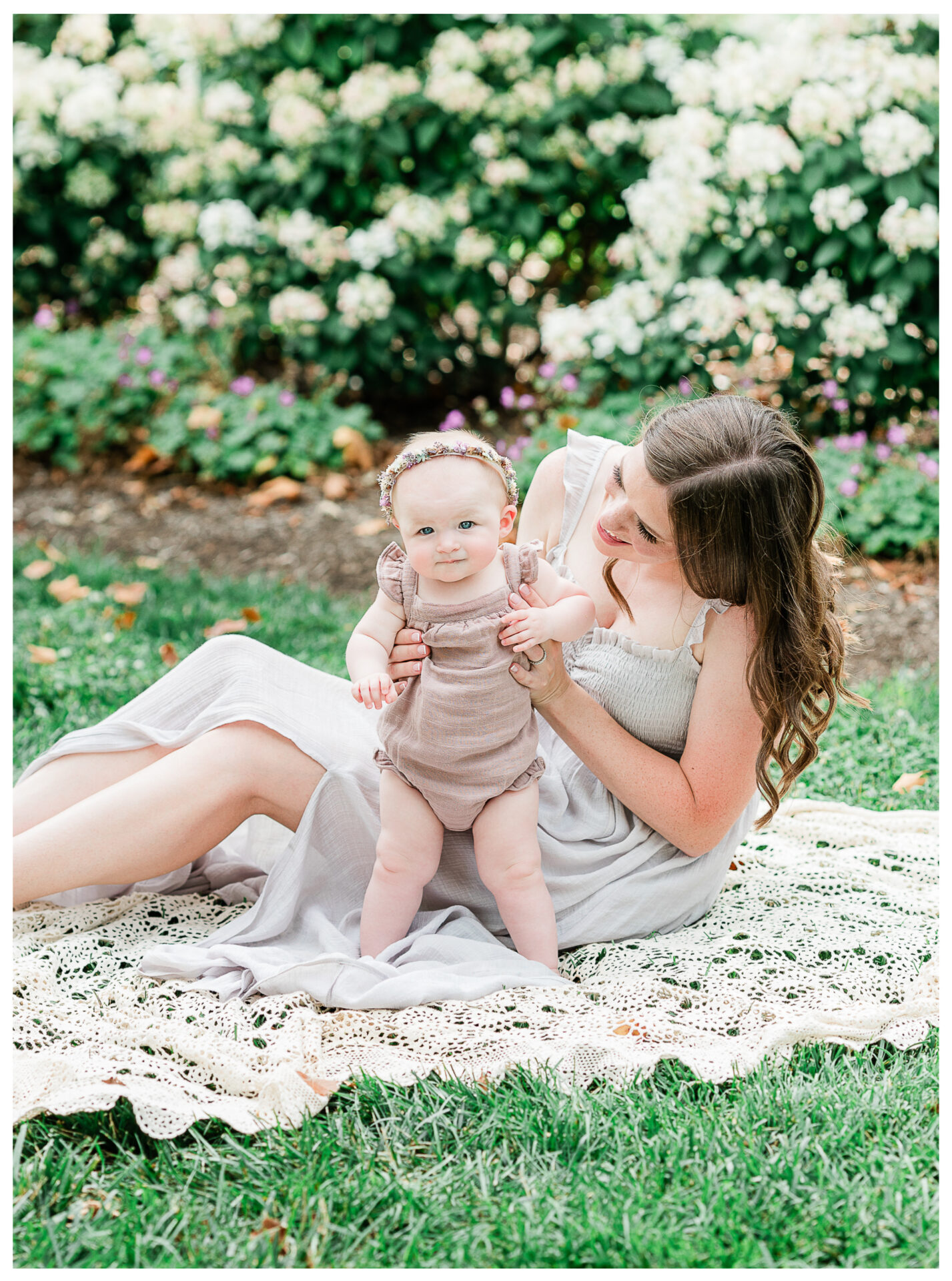 A mother helping her smiling baby girl stand on a lace blanket.