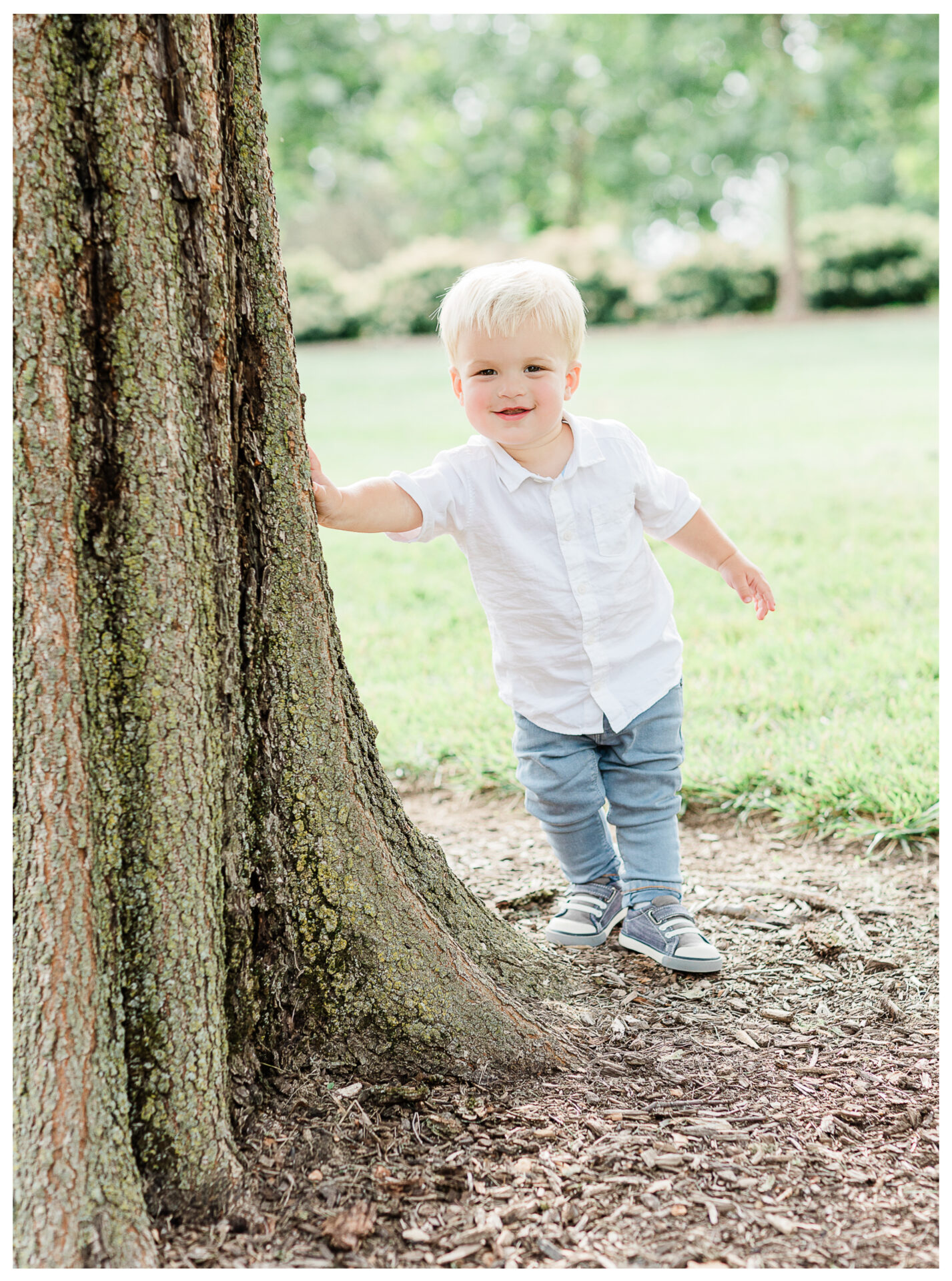 A toddler son smiling as he stands beside a large tree