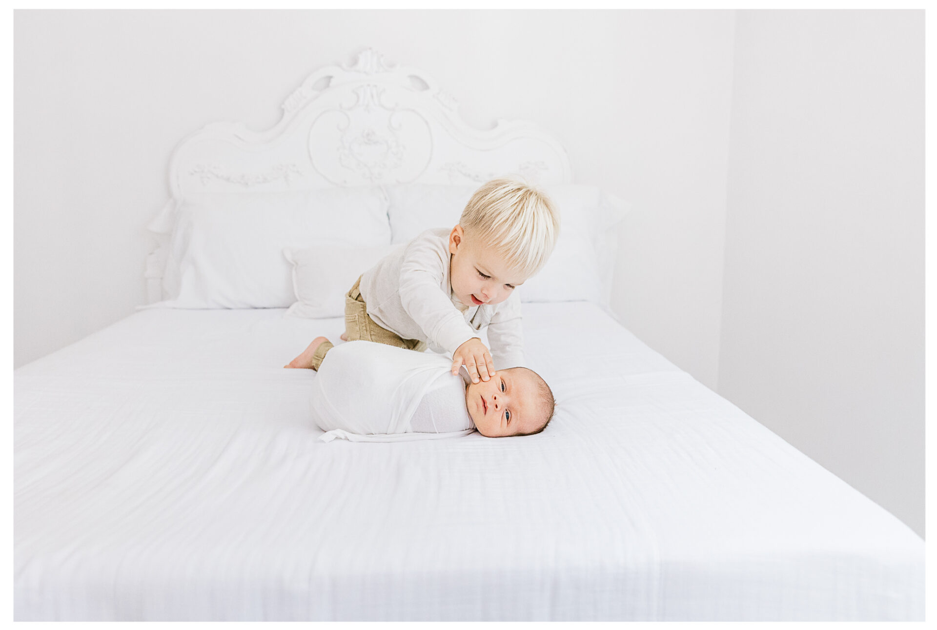 A young blonde toddler gently touches his swaddled baby brother who lies on a large white bed in a bright, airy room with white bedding and an ornate headboard.