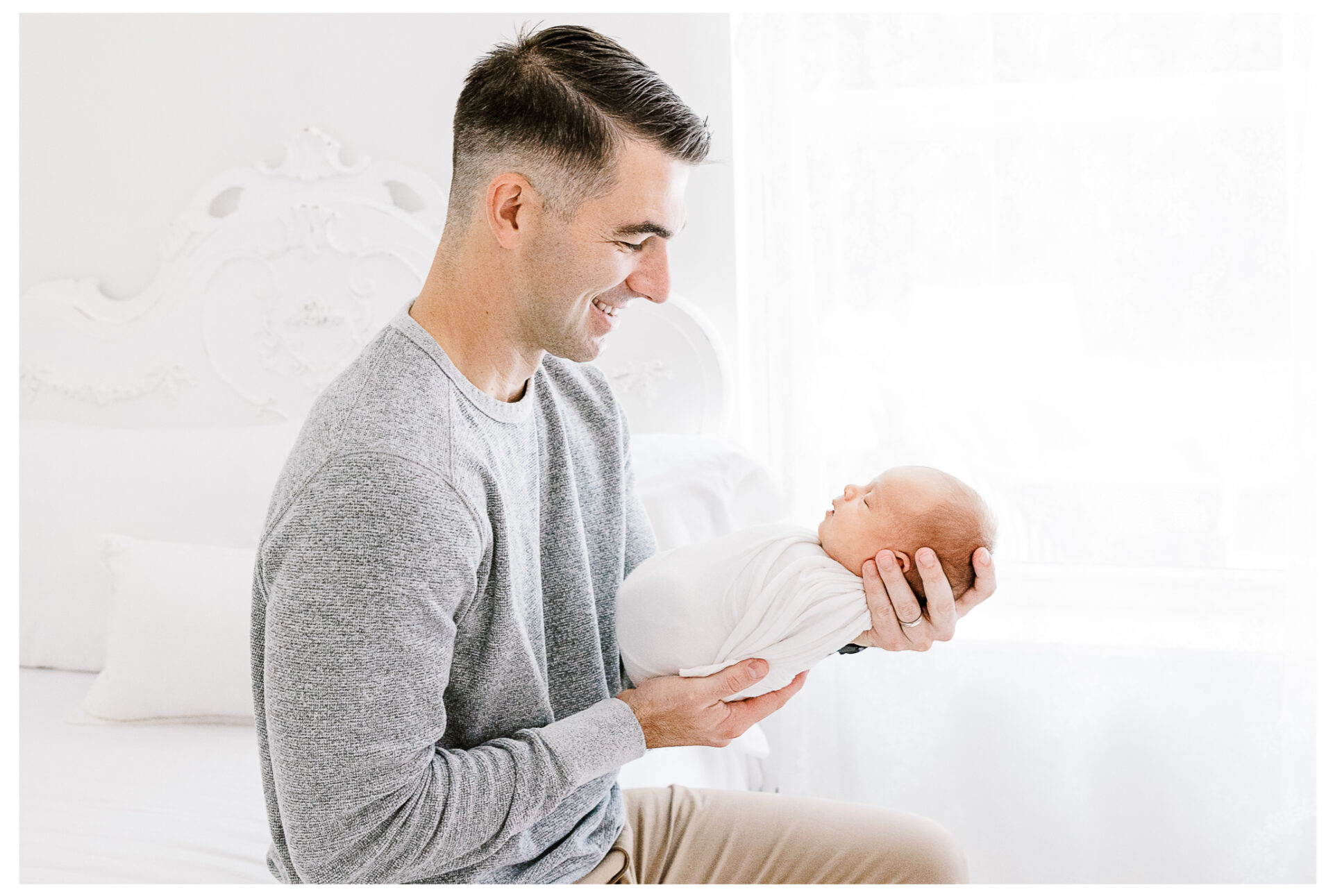 A father smiles while gently cradling his swaddled newborn son in his hands, sitting on a white bed in a bright, softly lit room.