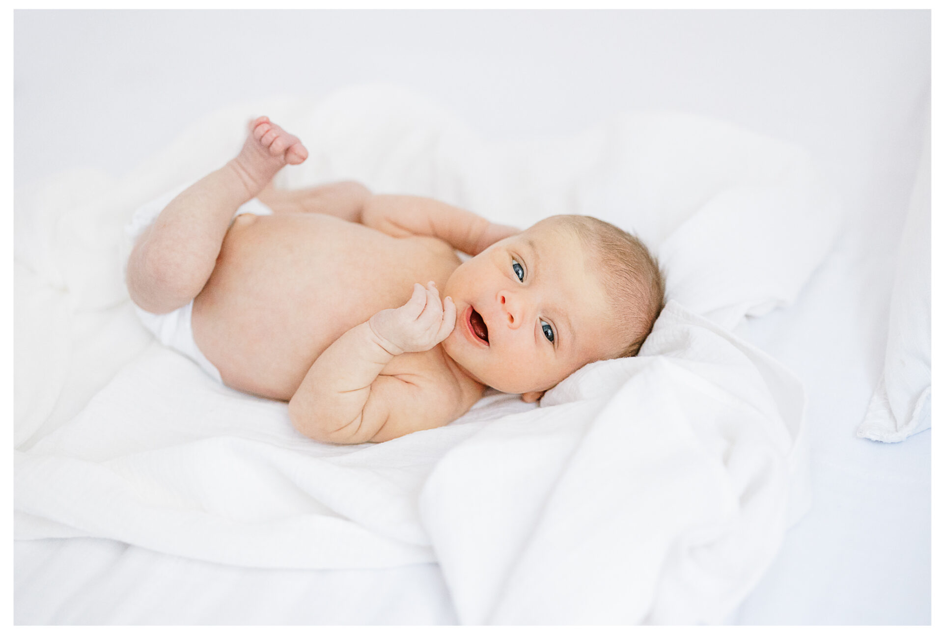 A newborn baby lies on a soft white blanket, looking up with wide eyes and mouth slightly open. The baby is wearing a diaper and appears relaxed and comfortable.