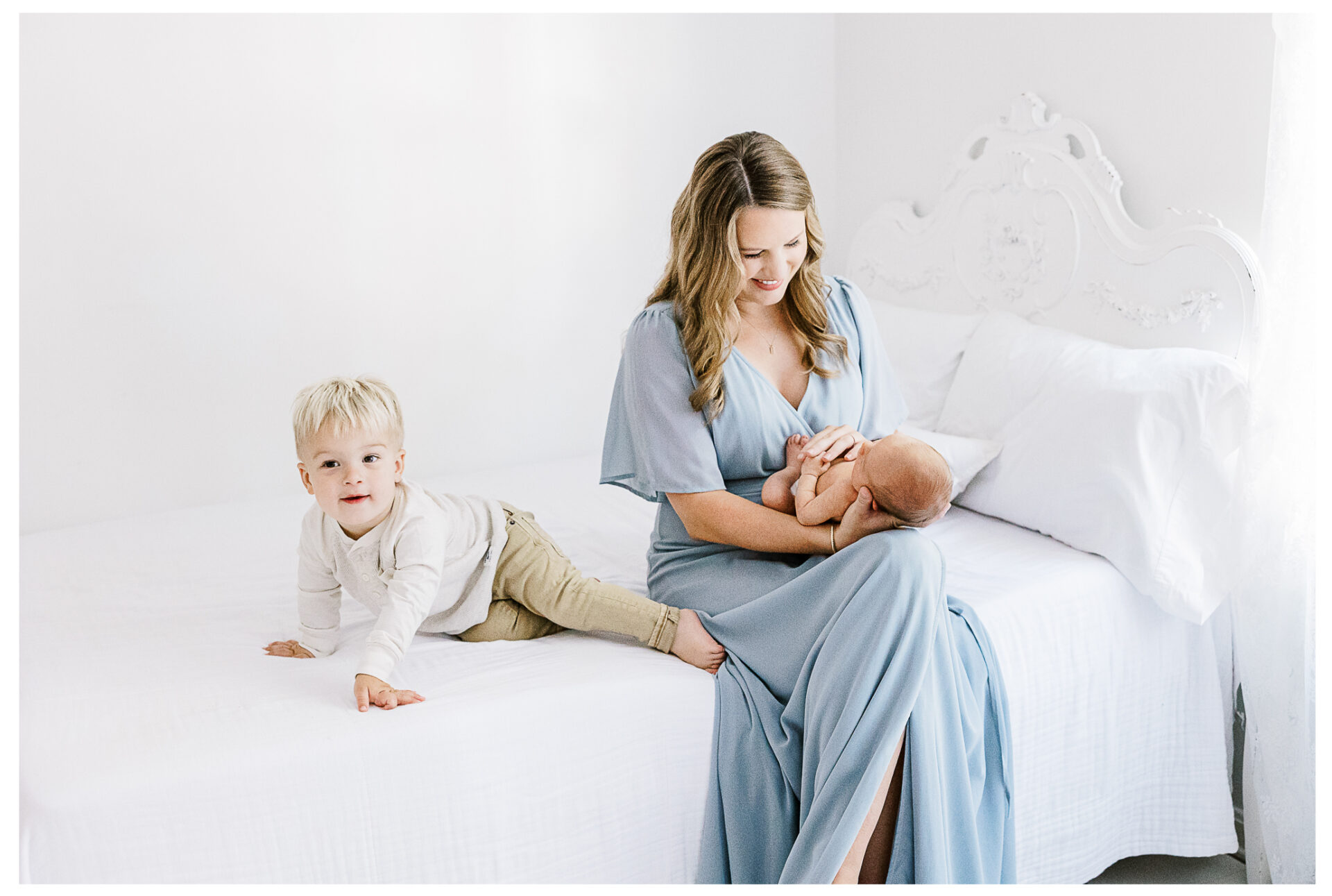 A mother in a light blue dress sits on a white bed holding her newborn, while her toddler son sits beside her smiling.