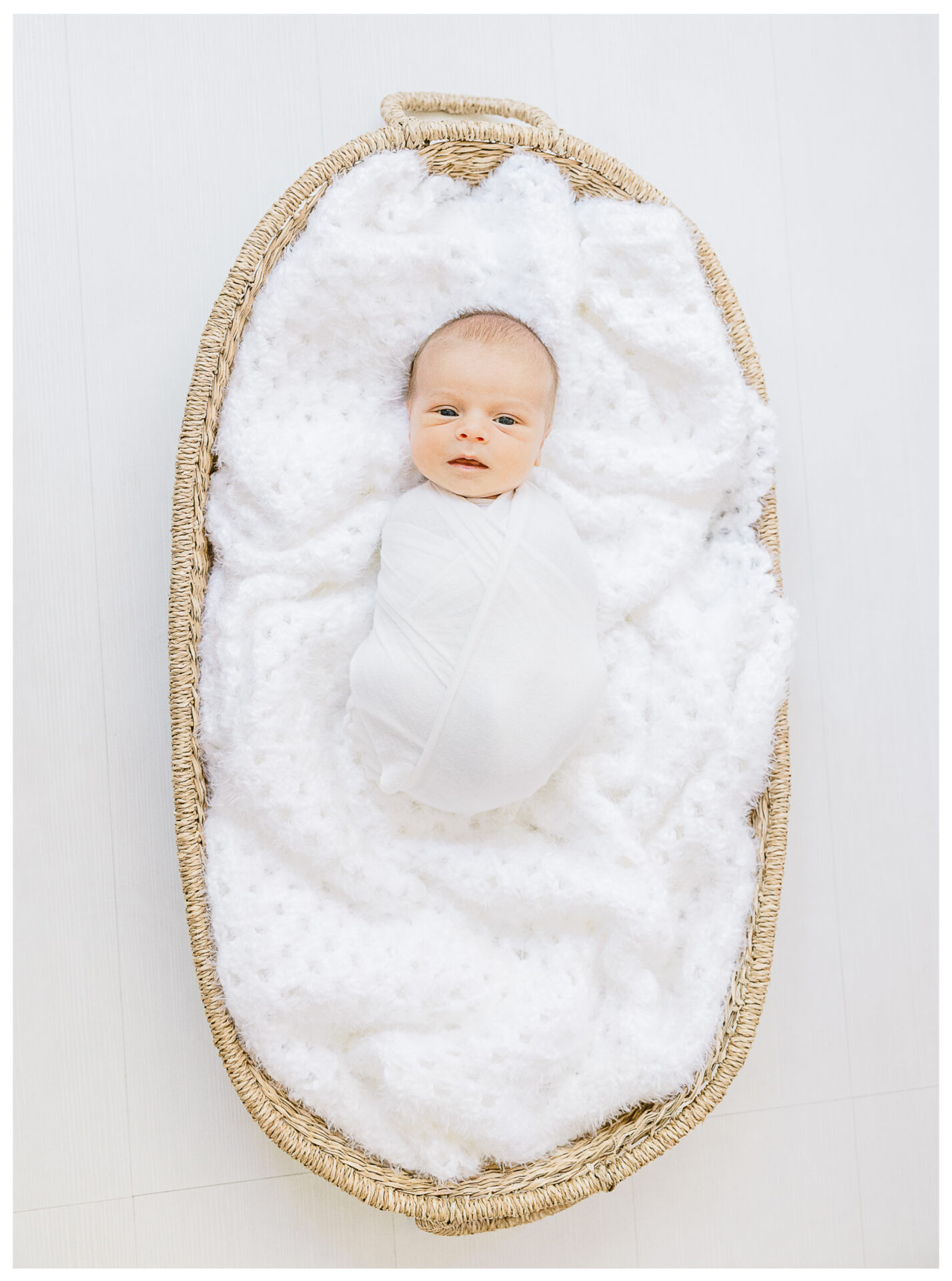 A newborn baby wrapped in a white blanket lies in a woven basket lined with a soft, textured white fabric, viewed from above on a light-colored floor.