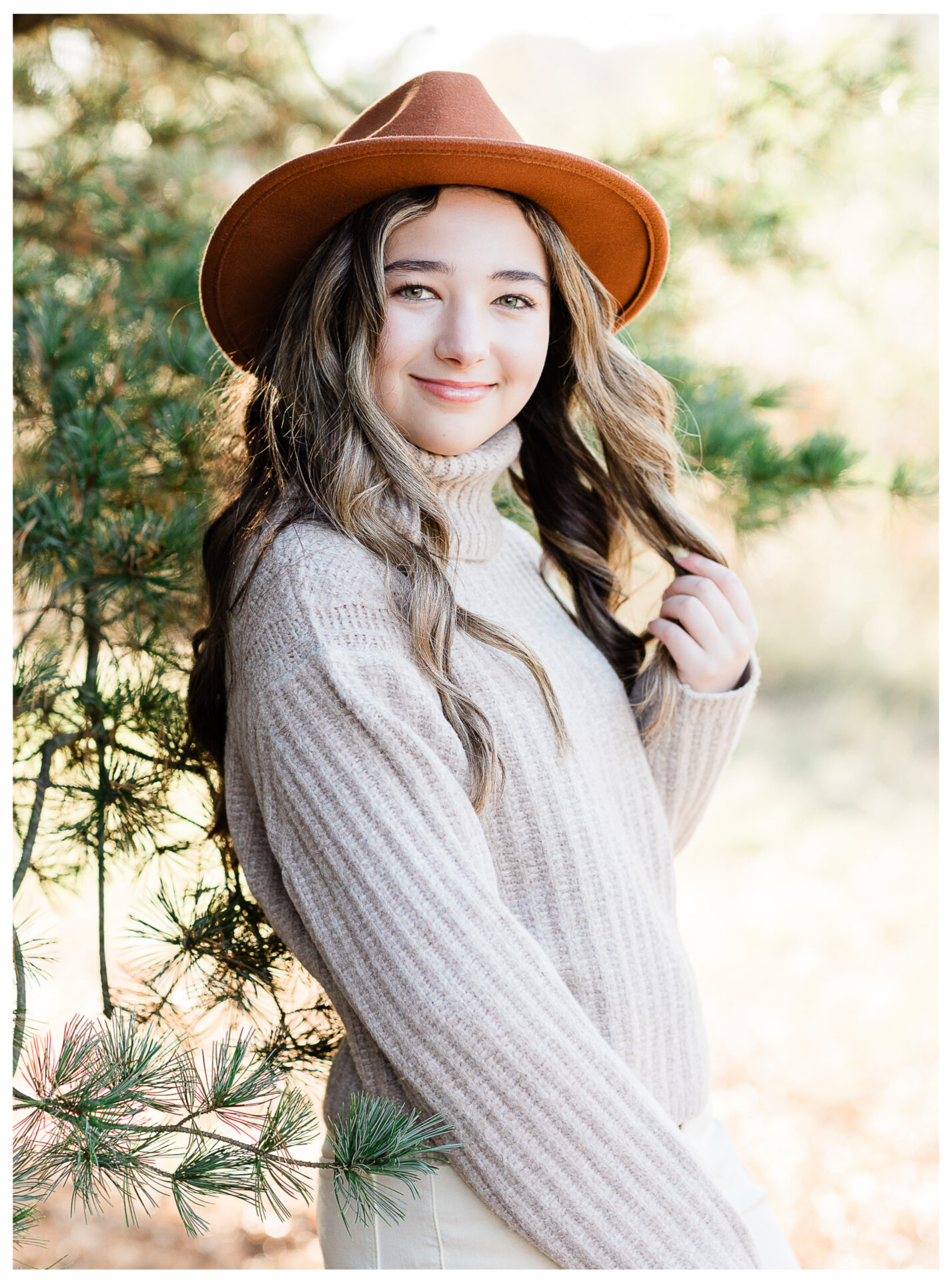 A preteen girl with long wavy hair, wearing a brown hat and beige turtleneck sweater, smiles while standing outdoors by a pine tree, with sunlight softly illuminating the background.
