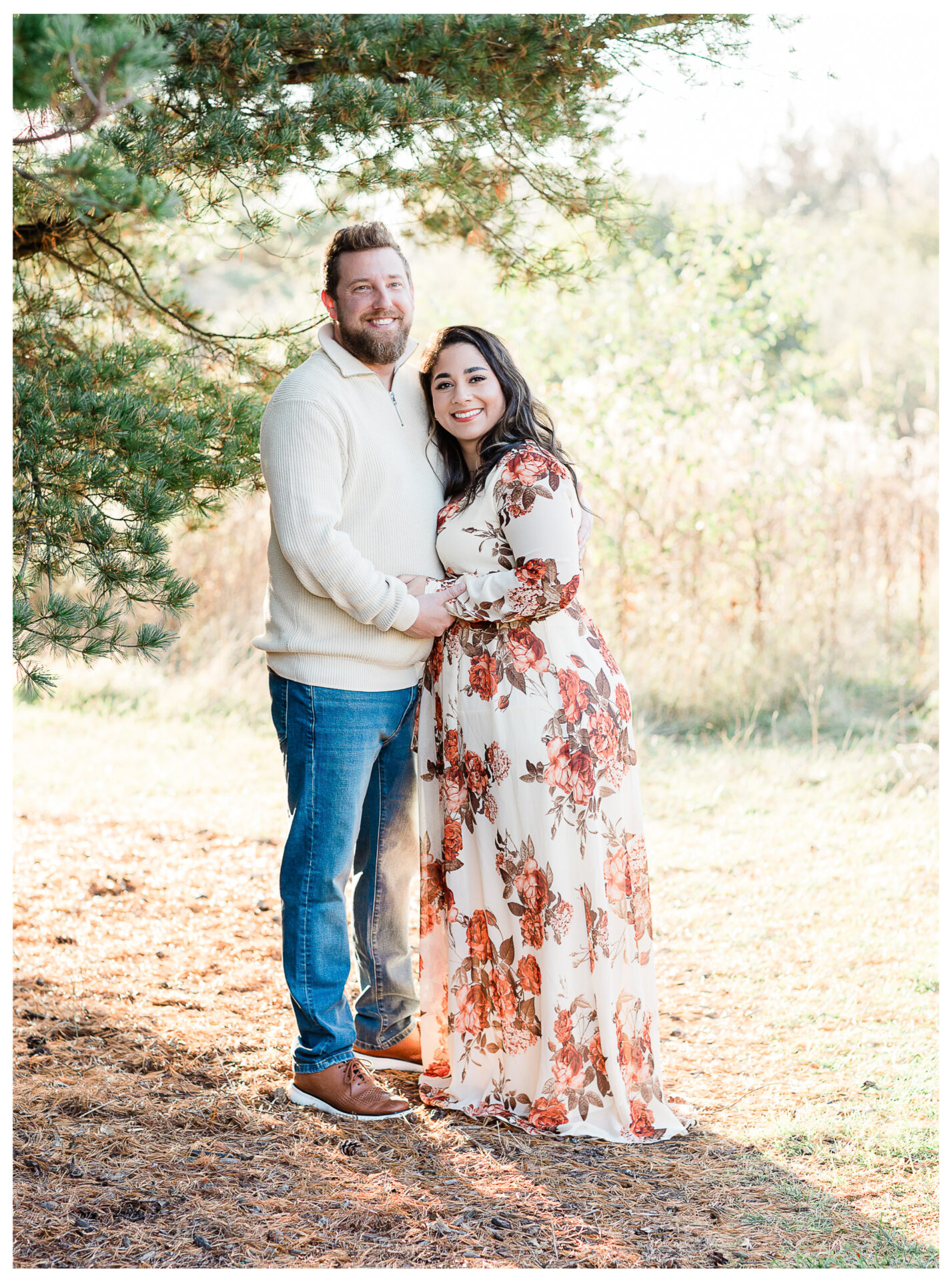 A smiling couple stands outdoors under a pine tree. The man wears a cream sweater and jeans, and the woman wears a long, floral dress. They are holding hands and posing together in the sunlight.