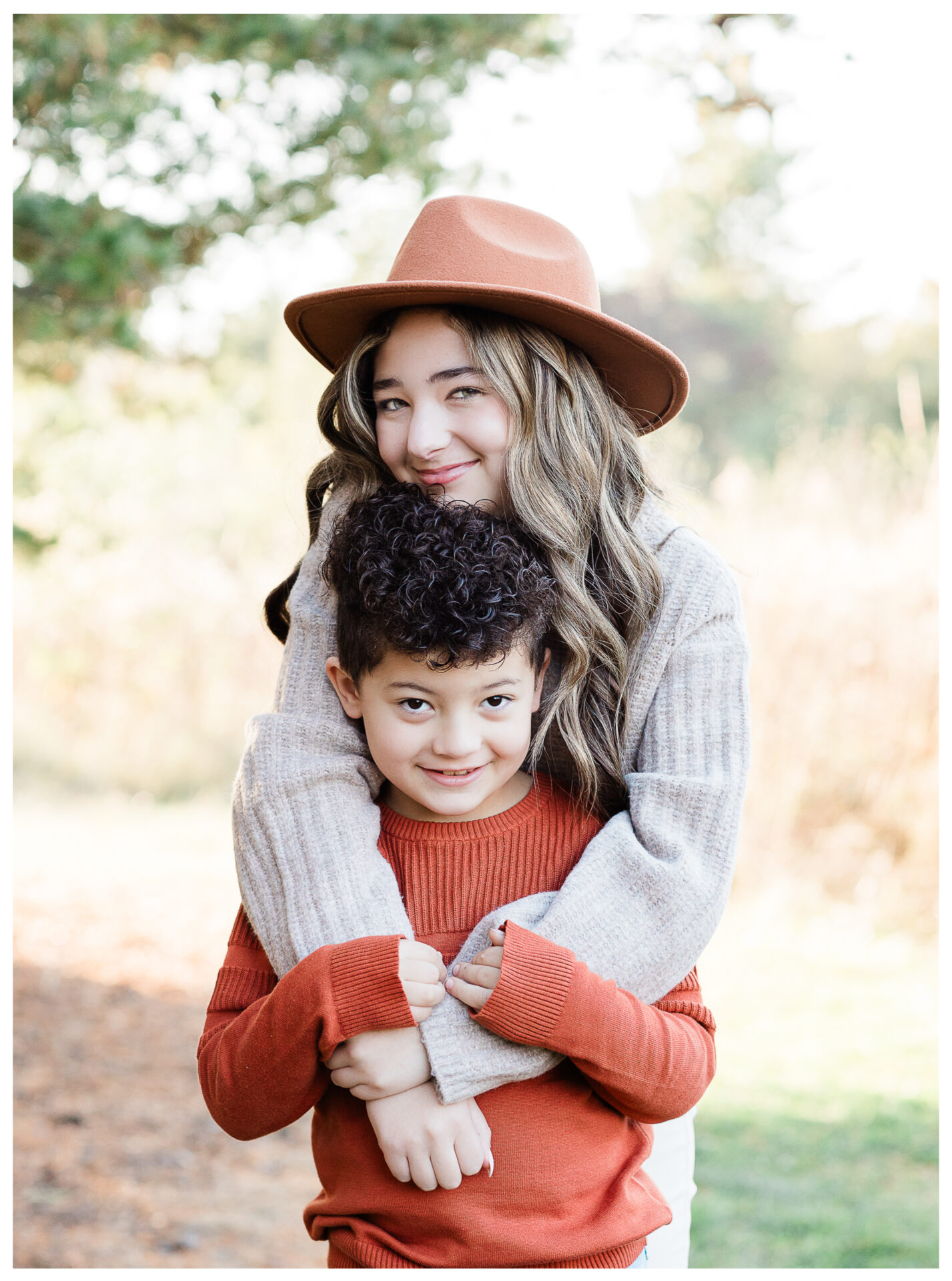 An older sister in a brown hat hugs her smiling young brother from behind. Both wear cozy sweaters in earth tones, and they stand outdoors surrounded by fall trees and soft sunlight.
