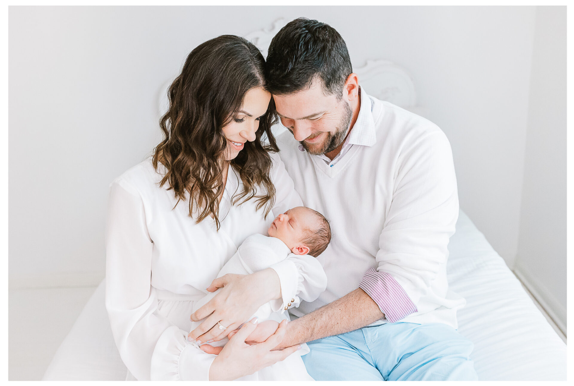 A husband and wife sitting on a white bed holding their newborn son between them.