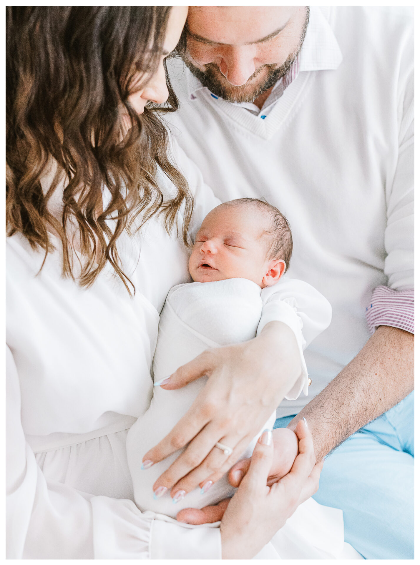 A husband and wife sitting on a white bed holding their newborn son between them.