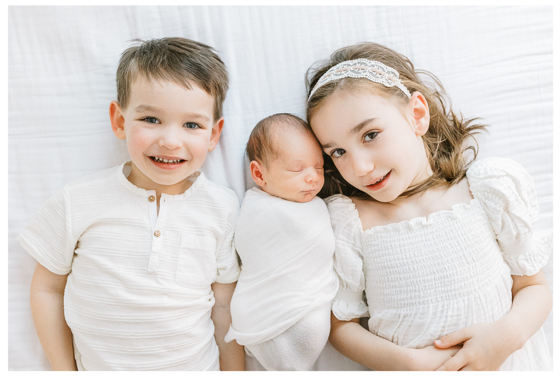 A young brother and sister smile broadly as they lie on their backs with their newborn brother asleep between them.