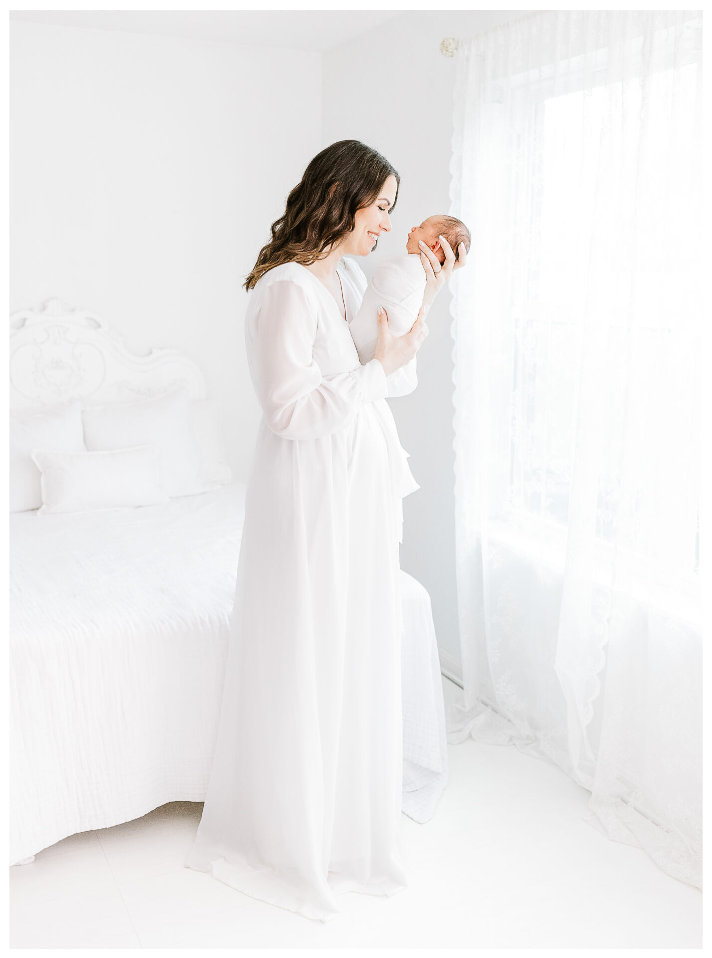 A mother smiling widely at her newborn baby while standing in the window light of a white room while wearing a long white gown.
