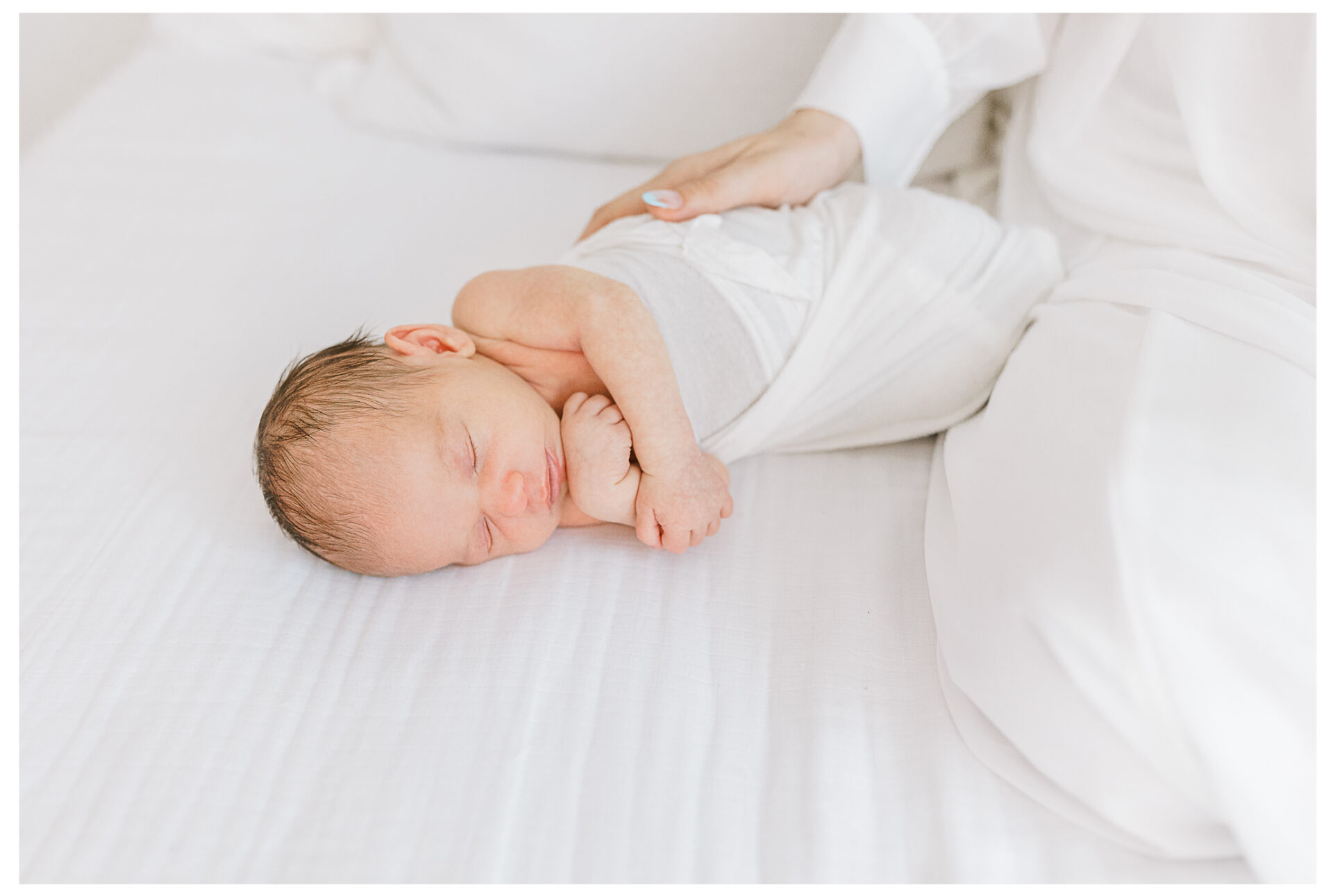 A mother's hand gently placed on her peacefully sleeping newborn son as he lies on a white bed.