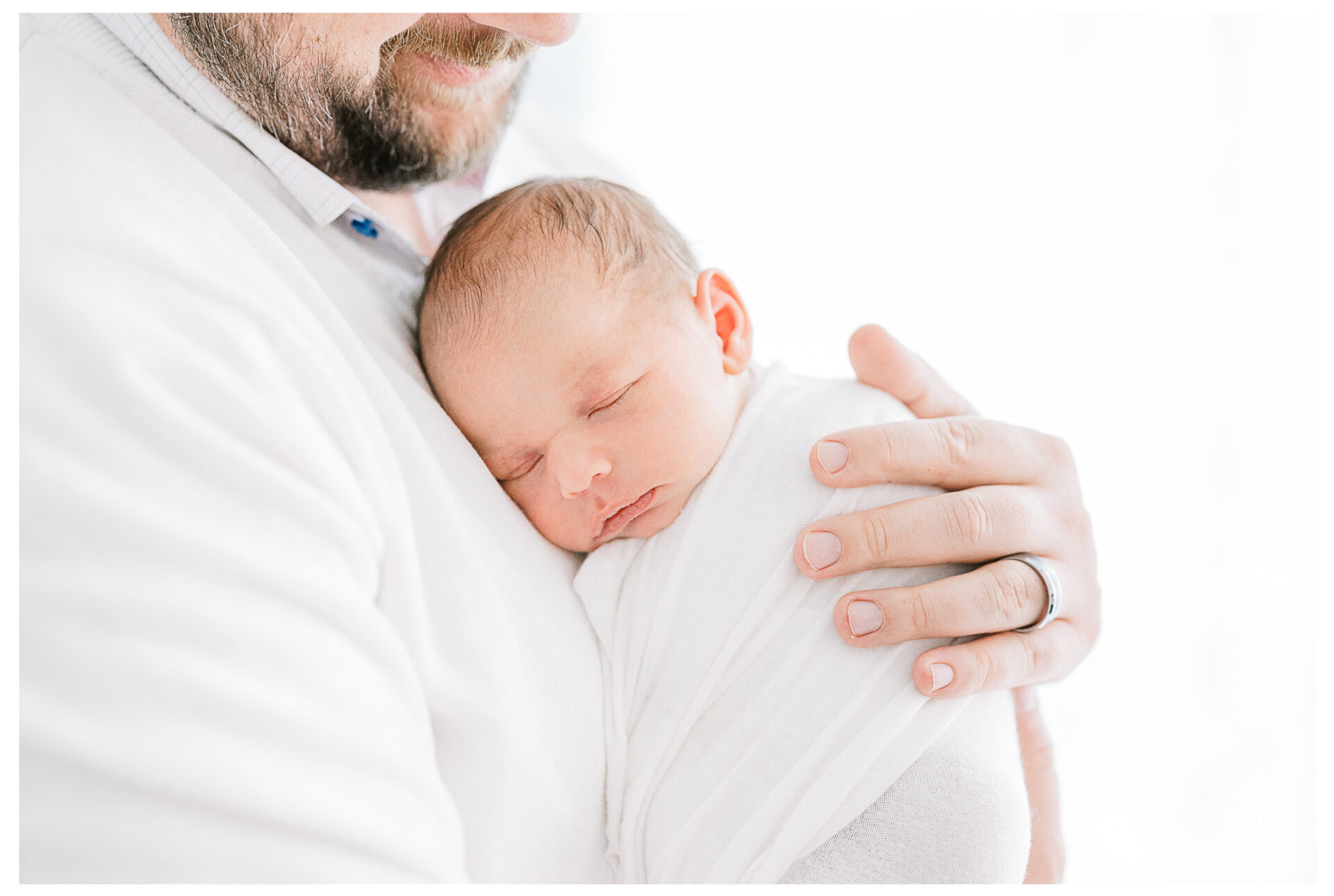 A close up of a swaddled sleeping newborn against his father's chest as the window light fills the background softly.