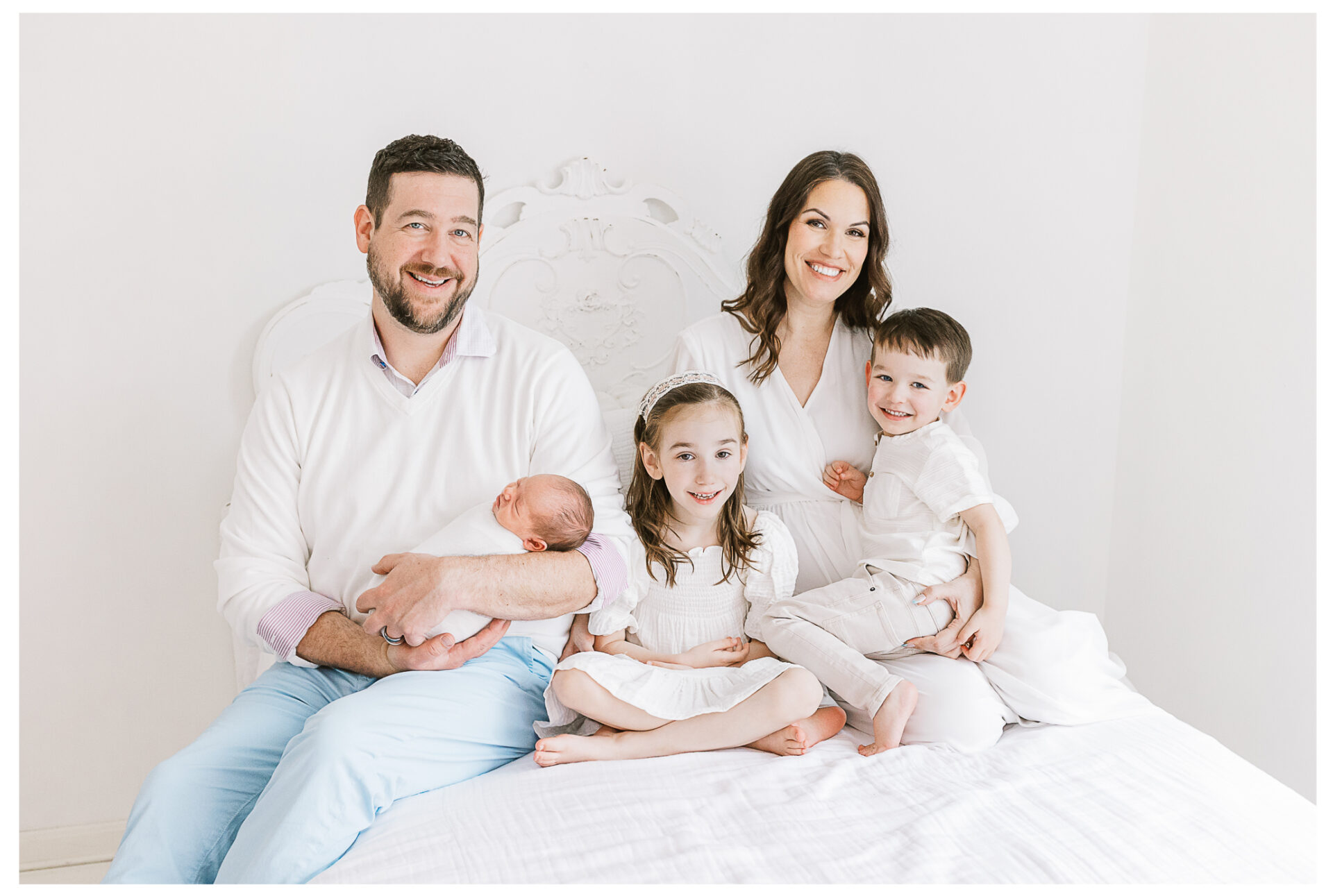 Parents sitting on a white bed smiling while snuggling their two young children and newborn baby.