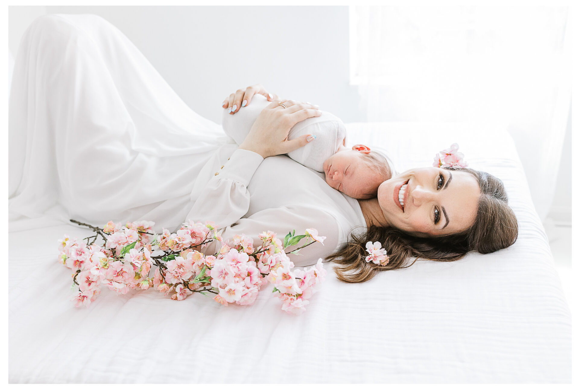 A smiling mother lying on a white bed surrounded by pink flowers with her newborn son asleep on her chest.