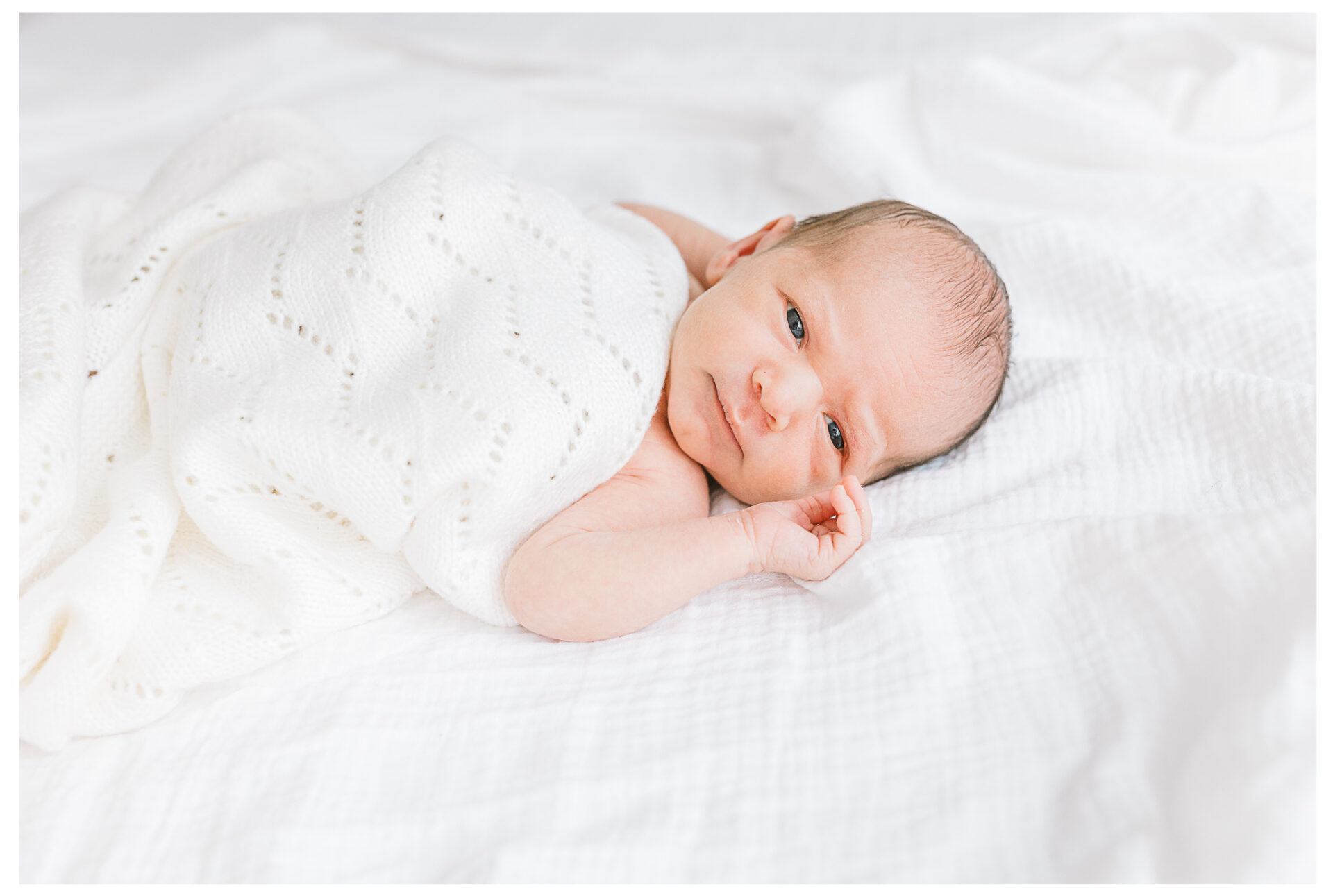 A newborn boy wrapped in a soft cream blanket with eyes wide open as he lies on a white bed wit a little smile on his face.