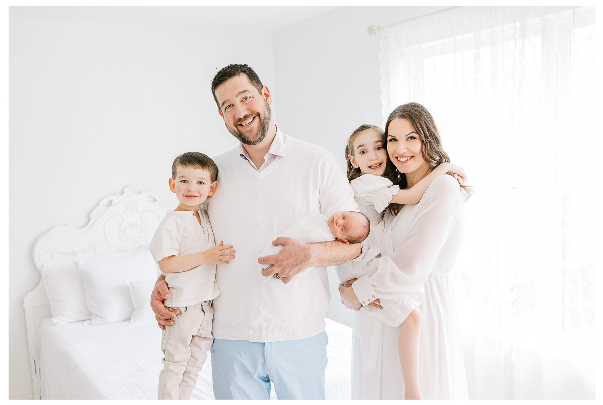 A family of five standing together beside a bright window as they smile and hold their newborn baby. Each member is wearing light clothing.