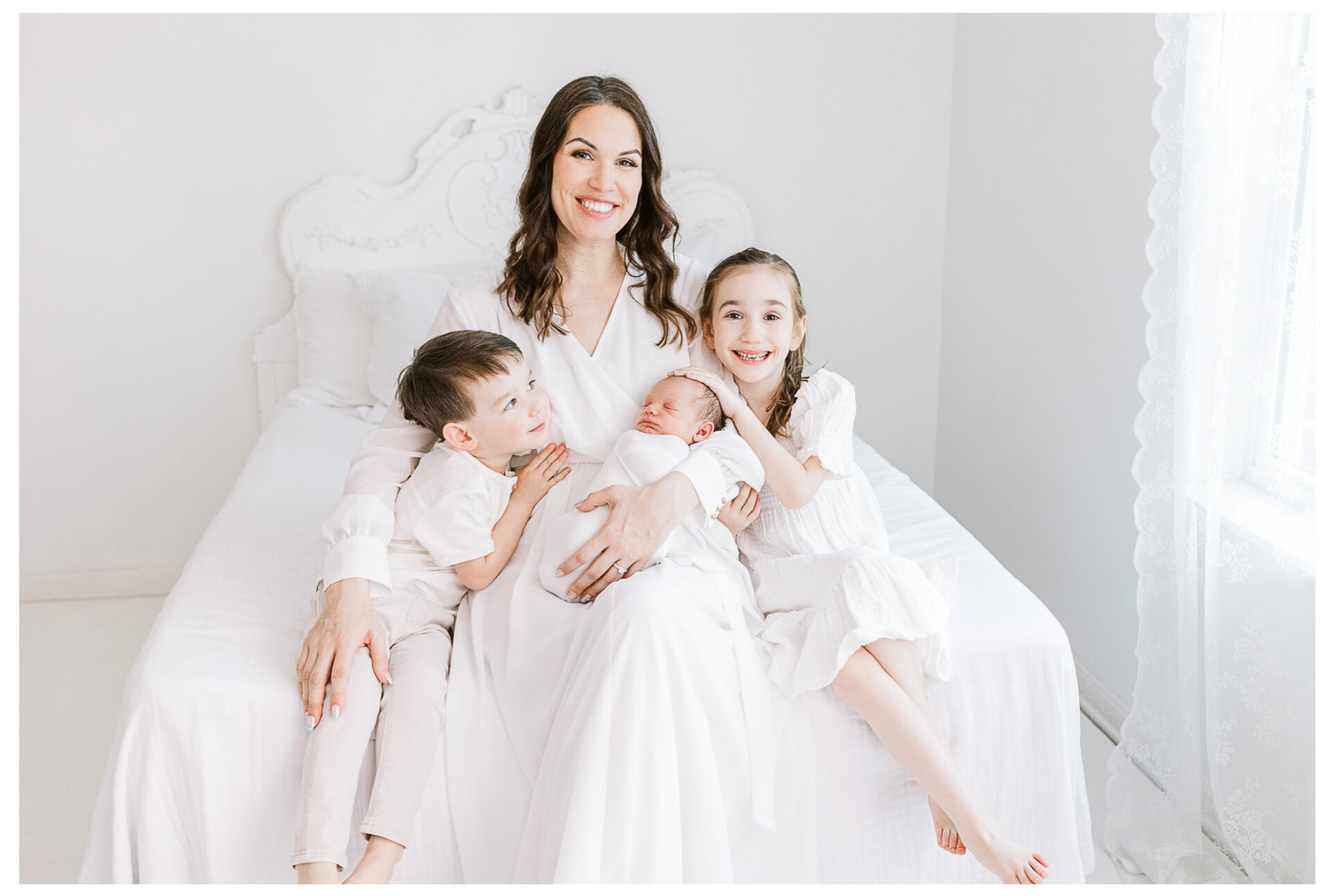 A mother holding her newborn baby as she smiles beside her other two young children. Everyone is wearing white as they sit on a white bed beside the window light.