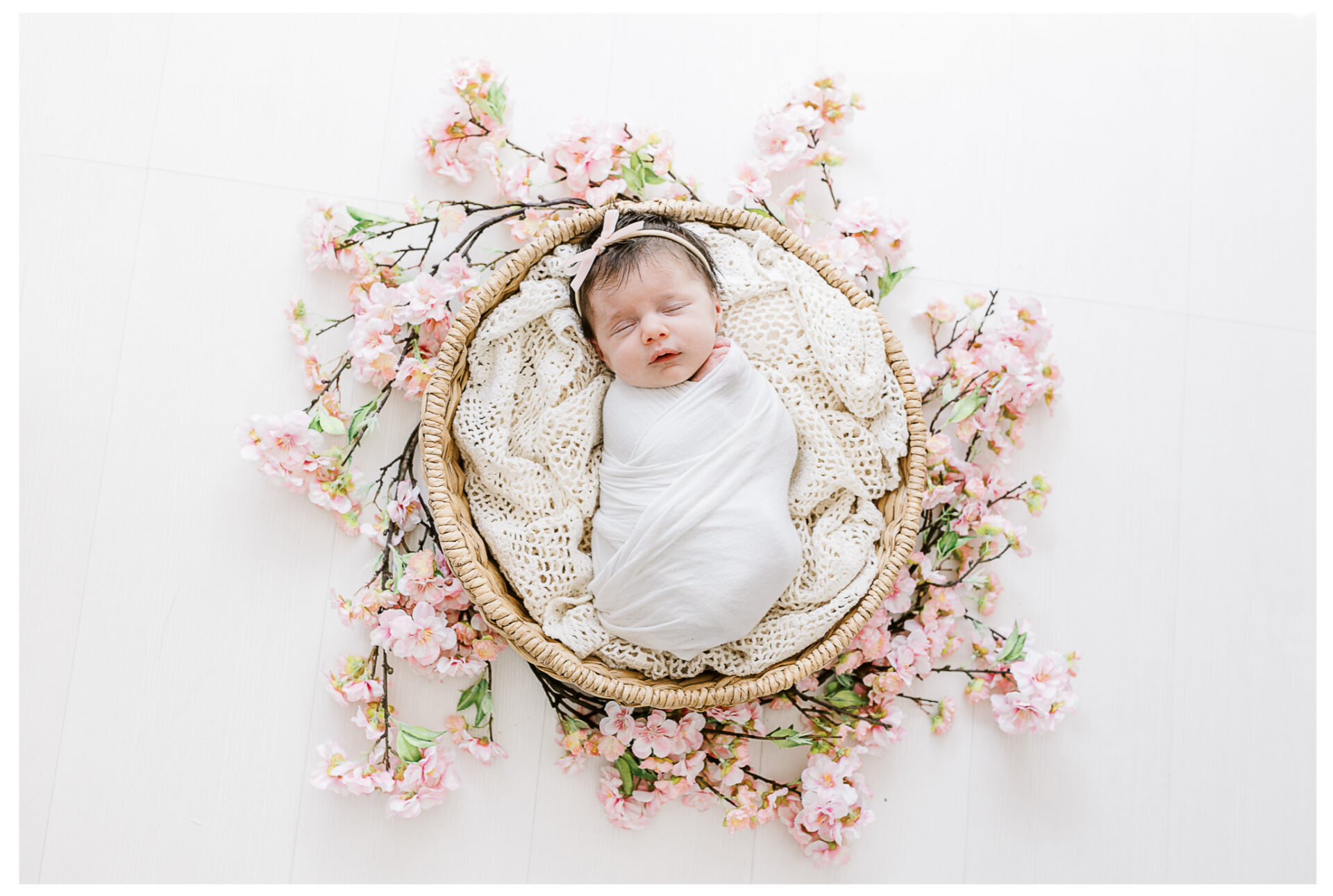 A newborn baby wrapped in a white blanket sleeps in a round wicker basket lined with a knitted cream blanket, surrounded by pink cherry blossom flowers on a white background.