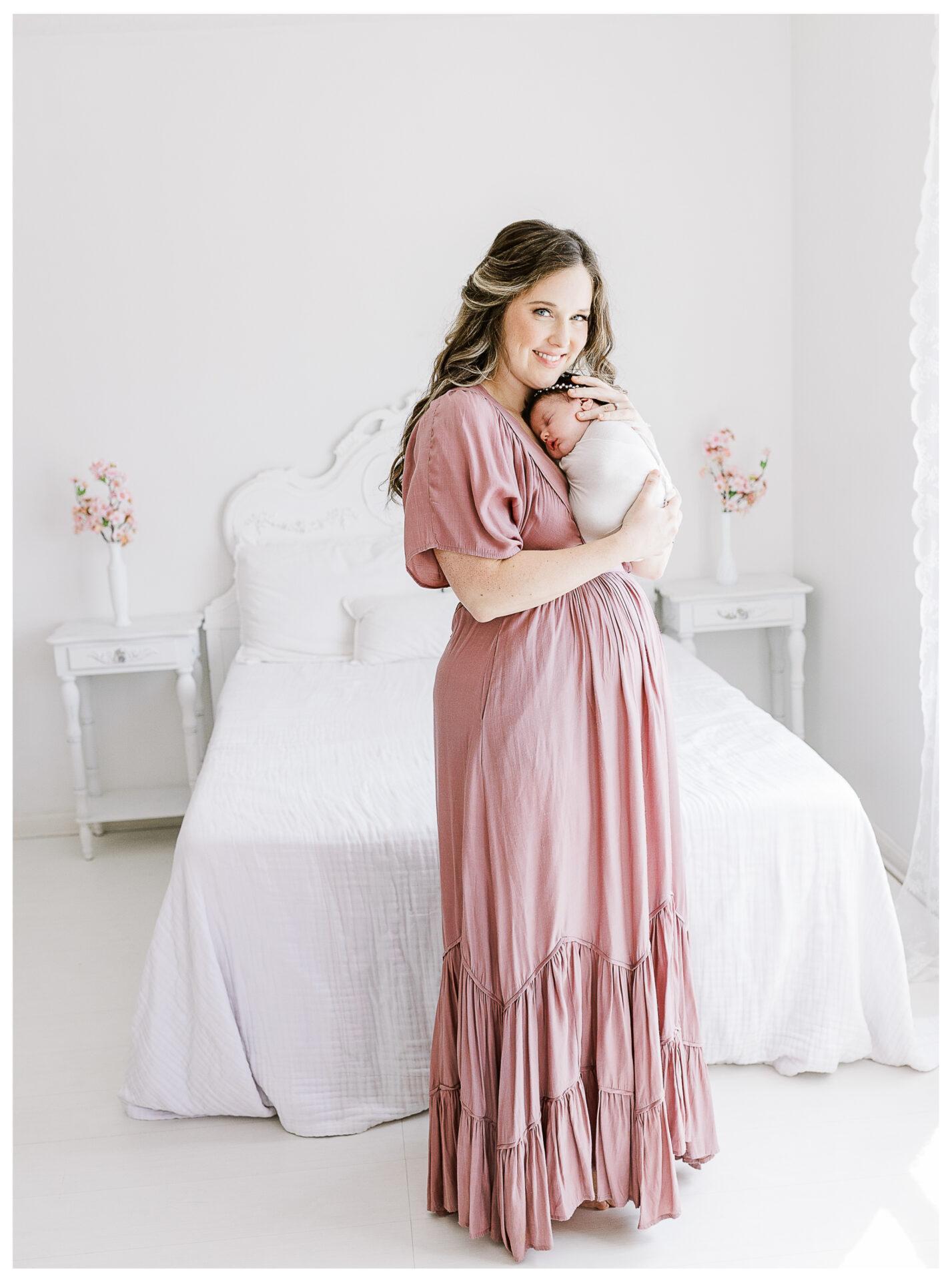 A mother in a long, pink dress stands in a bright, white bedroom, gently holding her newborn baby against her chest and smiling at the camera. The room has a white bed and two nightstands with pink flowers.
