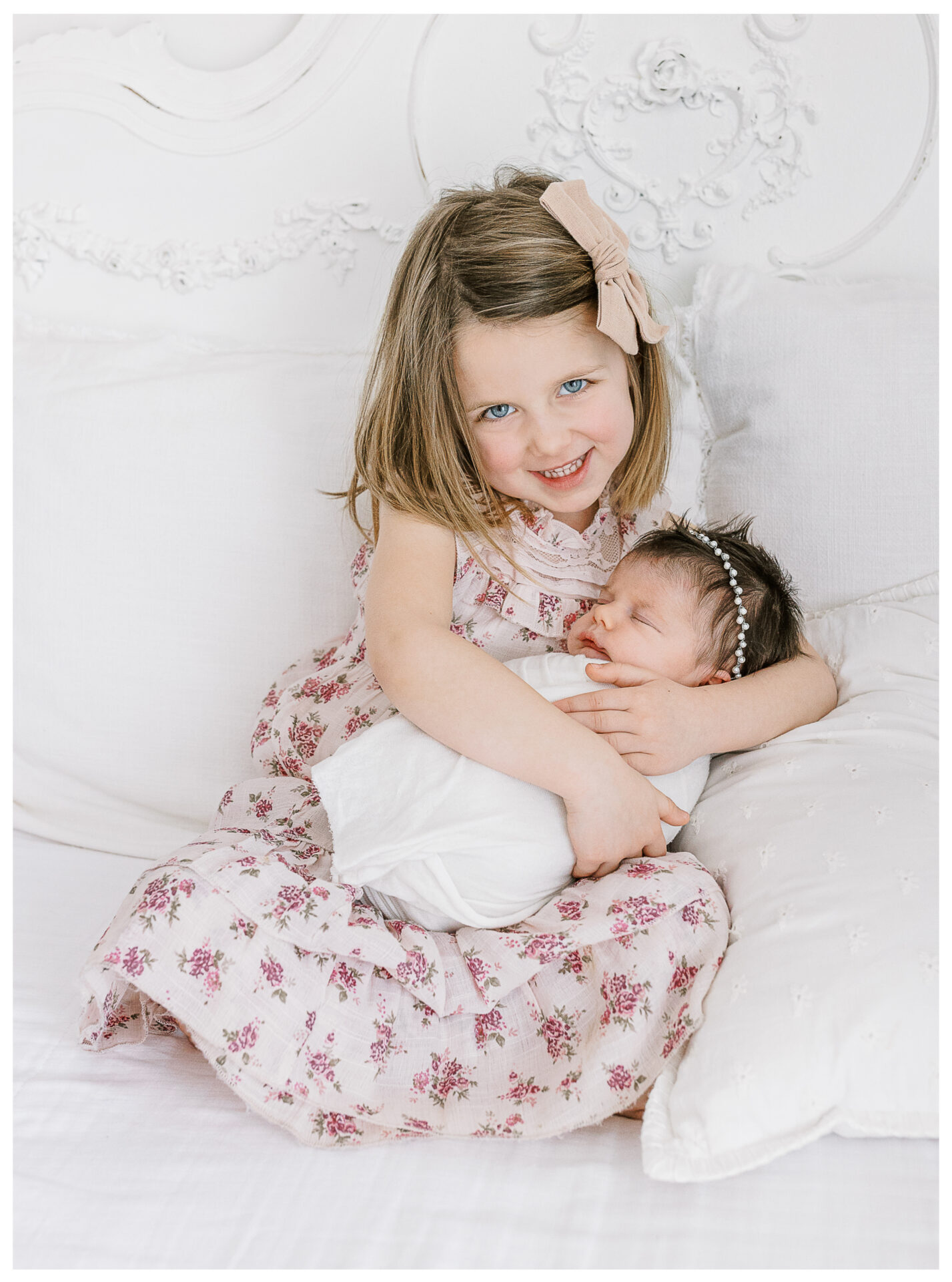 A young girl with shoulder-length hair and a pink bow smiles while sitting on a white bed, gently holding her swaddled newborn baby sister with dark hair and a beaded headband. Both wear light, floral-patterned clothing.