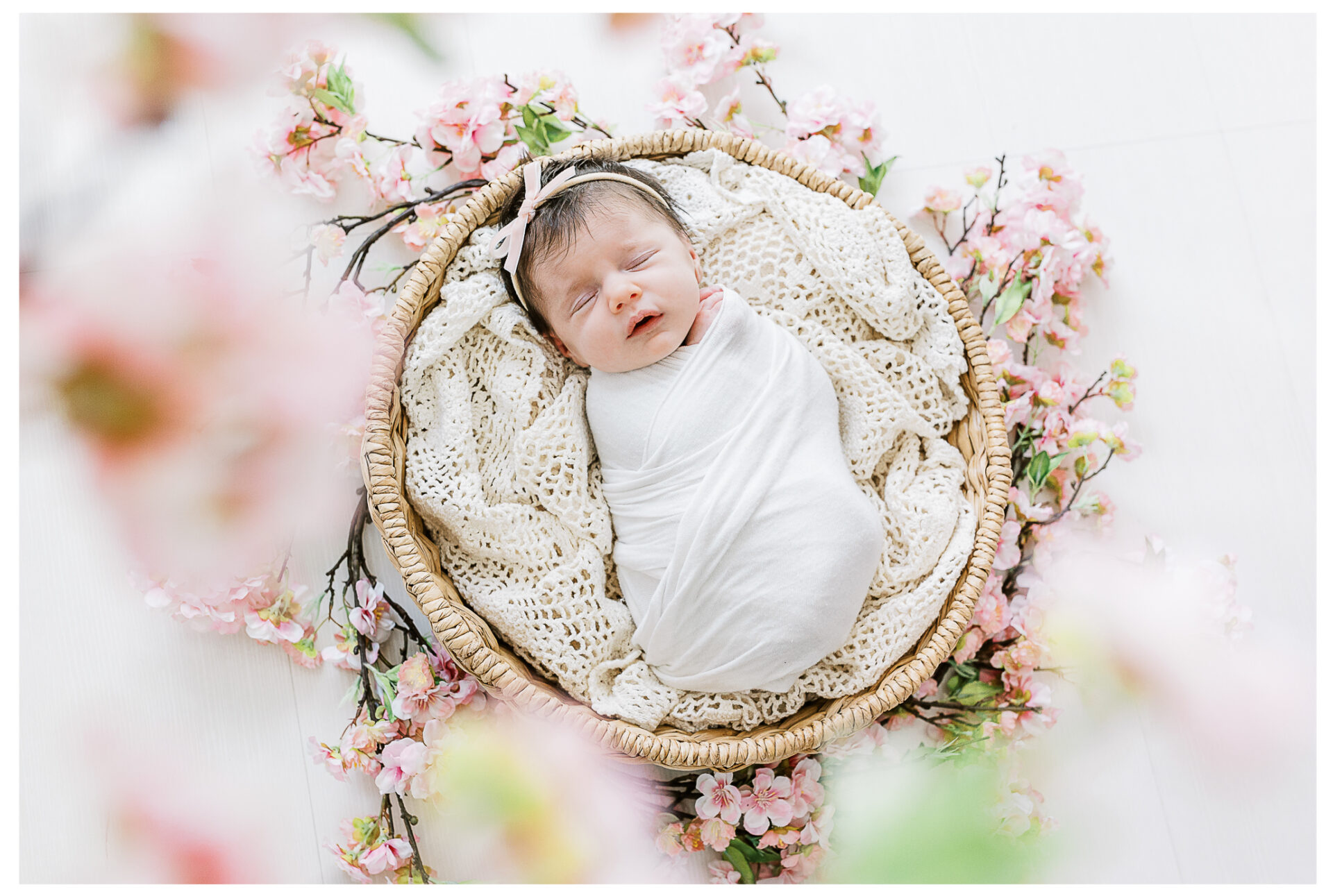A newborn baby wrapped in a white blanket sleeps peacefully in a round wicker basket, surrounded by soft pink flowers and a cream-colored knitted blanket.