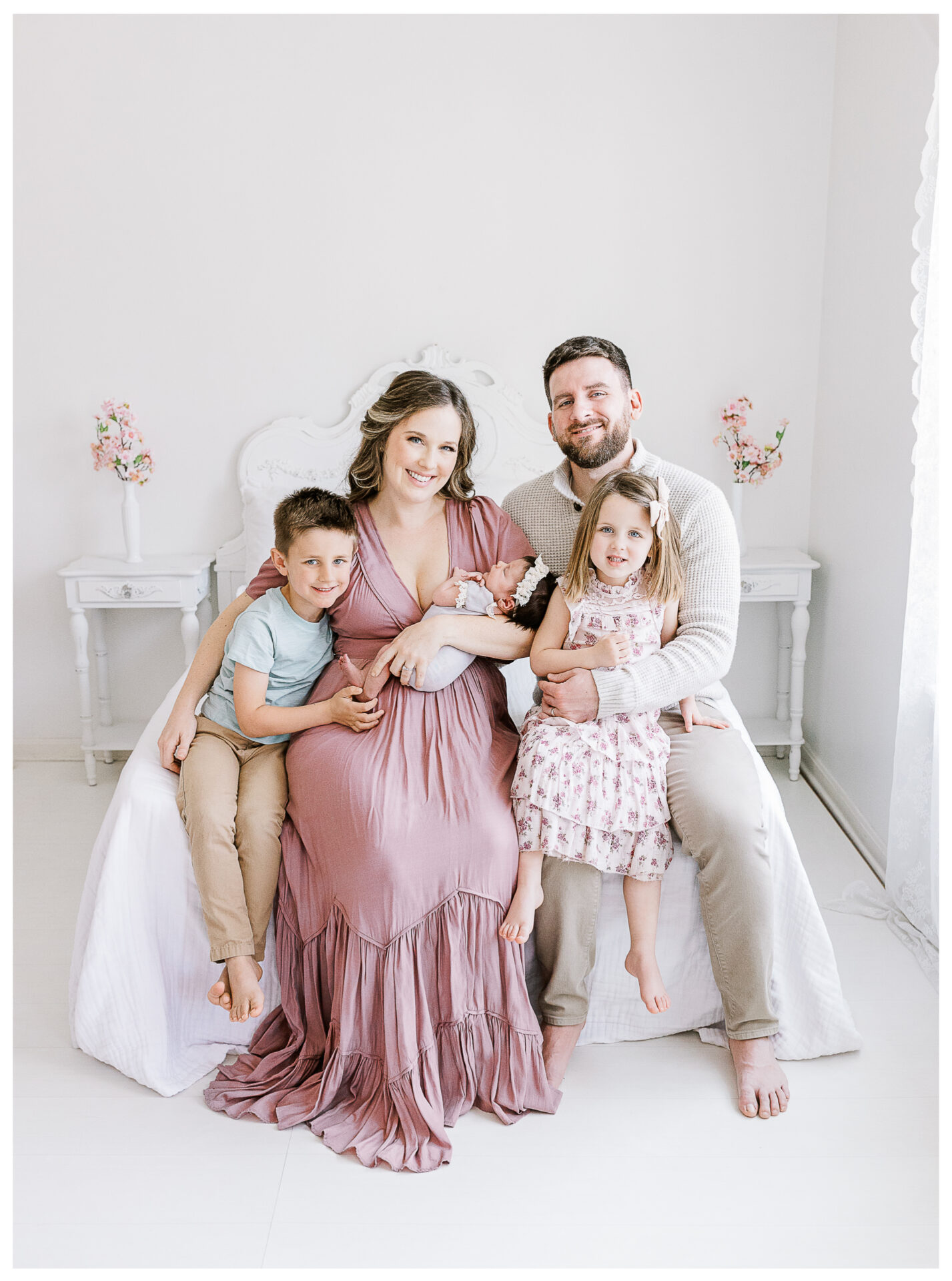 A smiling family of five sits on a white bed. The mother in a mauve dress holds their newborn, with a young boy on her left and a little girl in a floral dress on her right, sitting next to the father. The room is bright and airy.