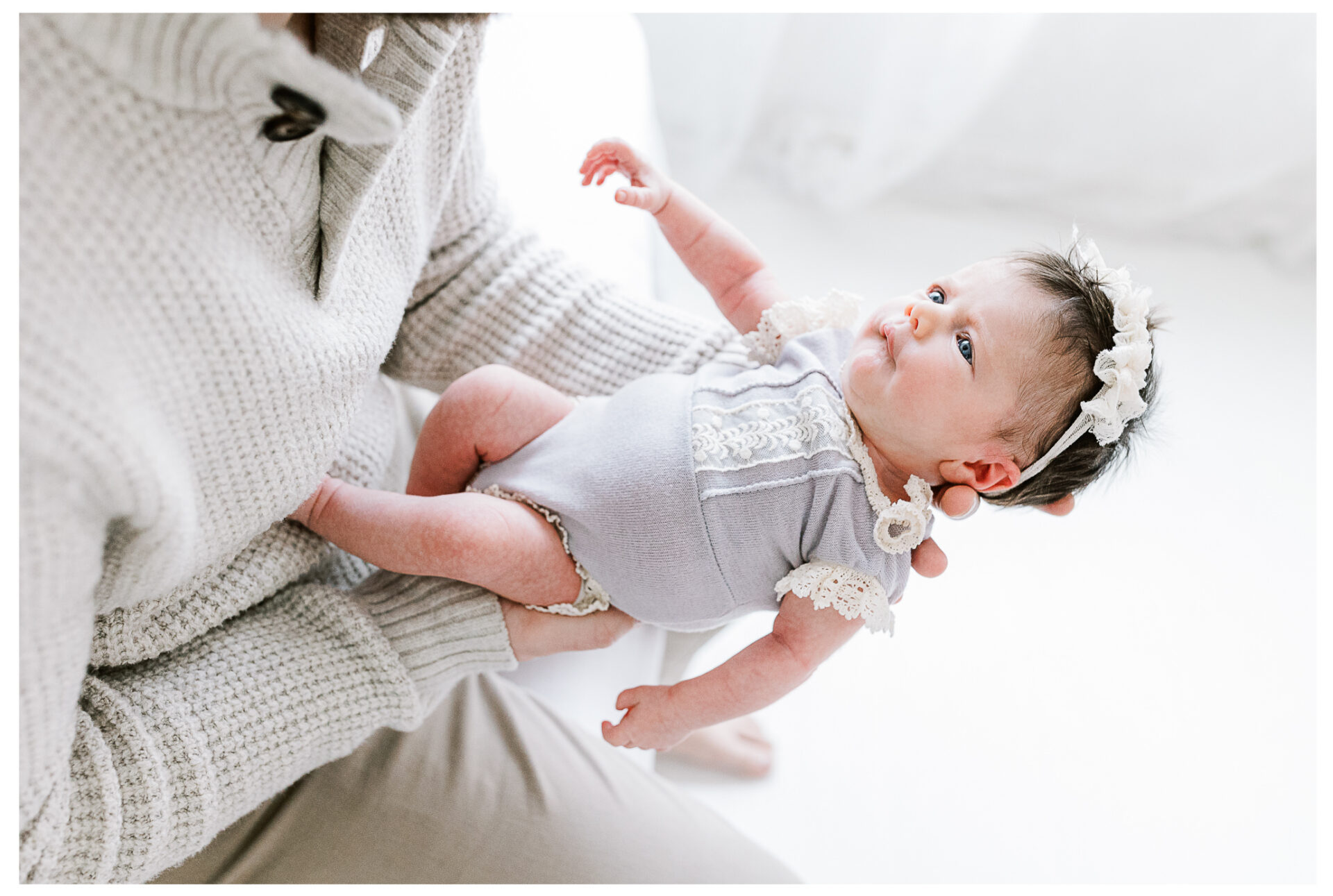 A father wearing a light knit sweater holds his baby dressed in a lavender romper with lace details and a floral headband during a sweet and sleepy newborn girl session in softly lit, white Centerville, Ohio setting.