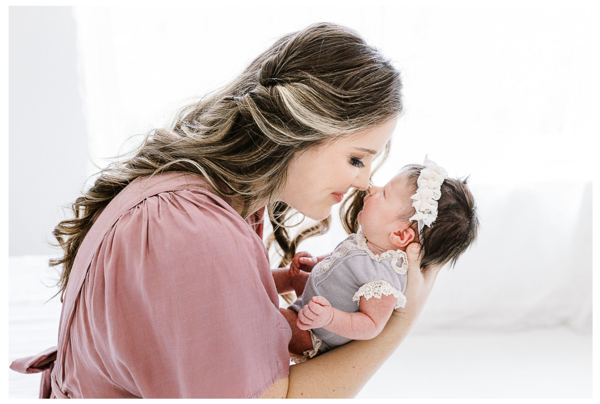 A mother with long, wavy hair in a pink dress gently holds her newborn baby girl. touching noses and smiling softly in a bright, airy room.