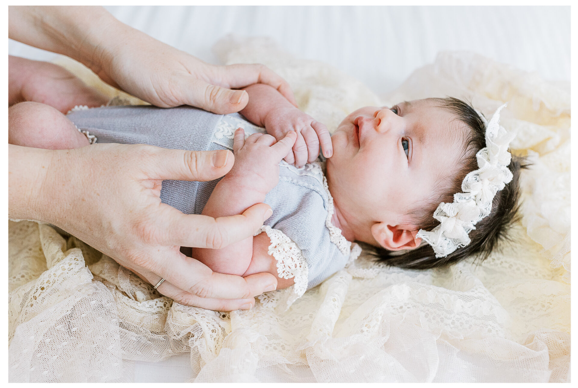 A newborn baby girl wearing a lace headband and lavender colored outfit lies on a soft, cream lace blanket while a mother gently holds the baby’s hands. The baby has a soft smile on her lips.