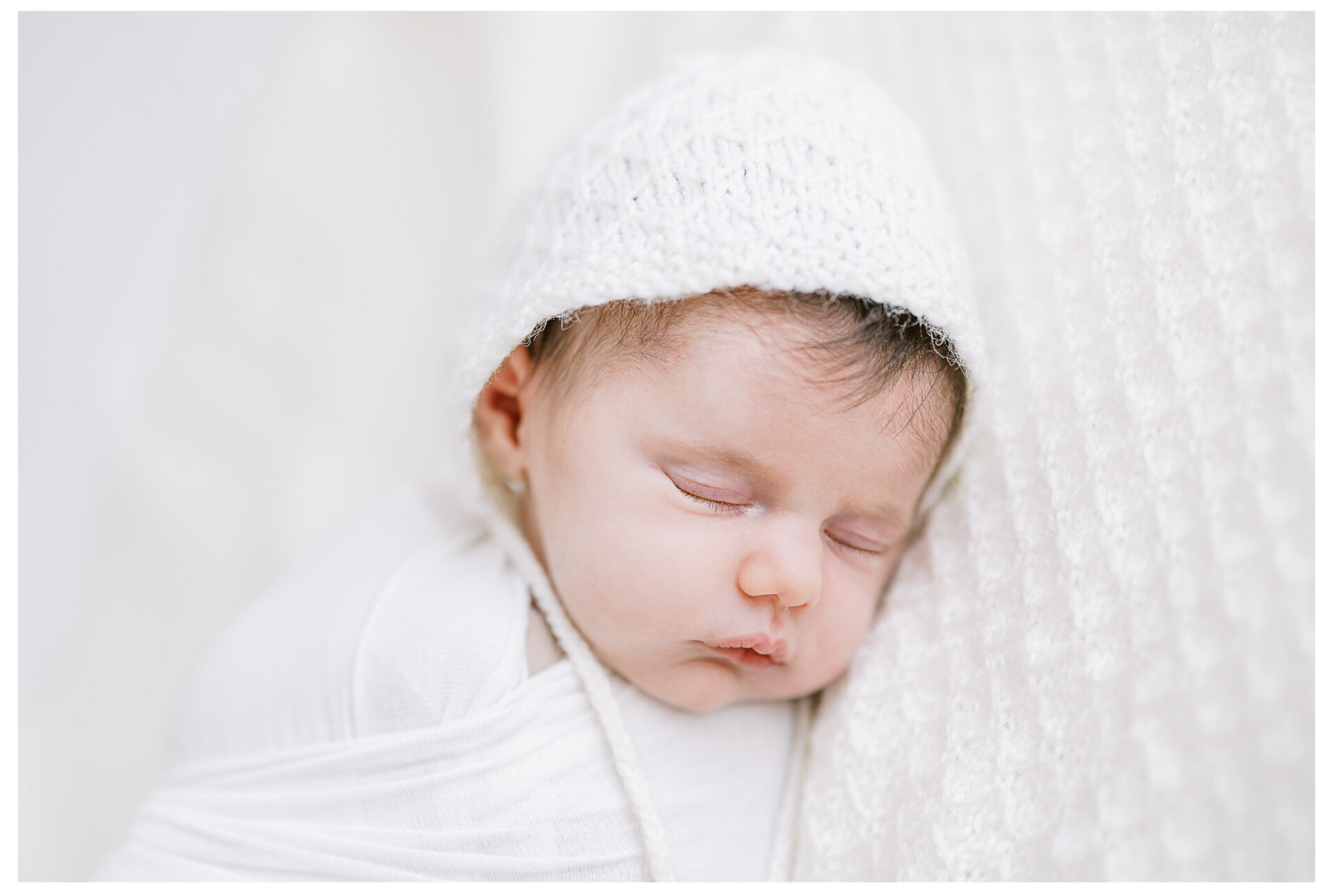A newborn baby sleeps peacefully, swaddled in a white blanket and wearing a knitted white bonnet, lying on a textured cream background. Winter Freire Photography | Centerville, Ohio sweet and sleepy newborn girl session