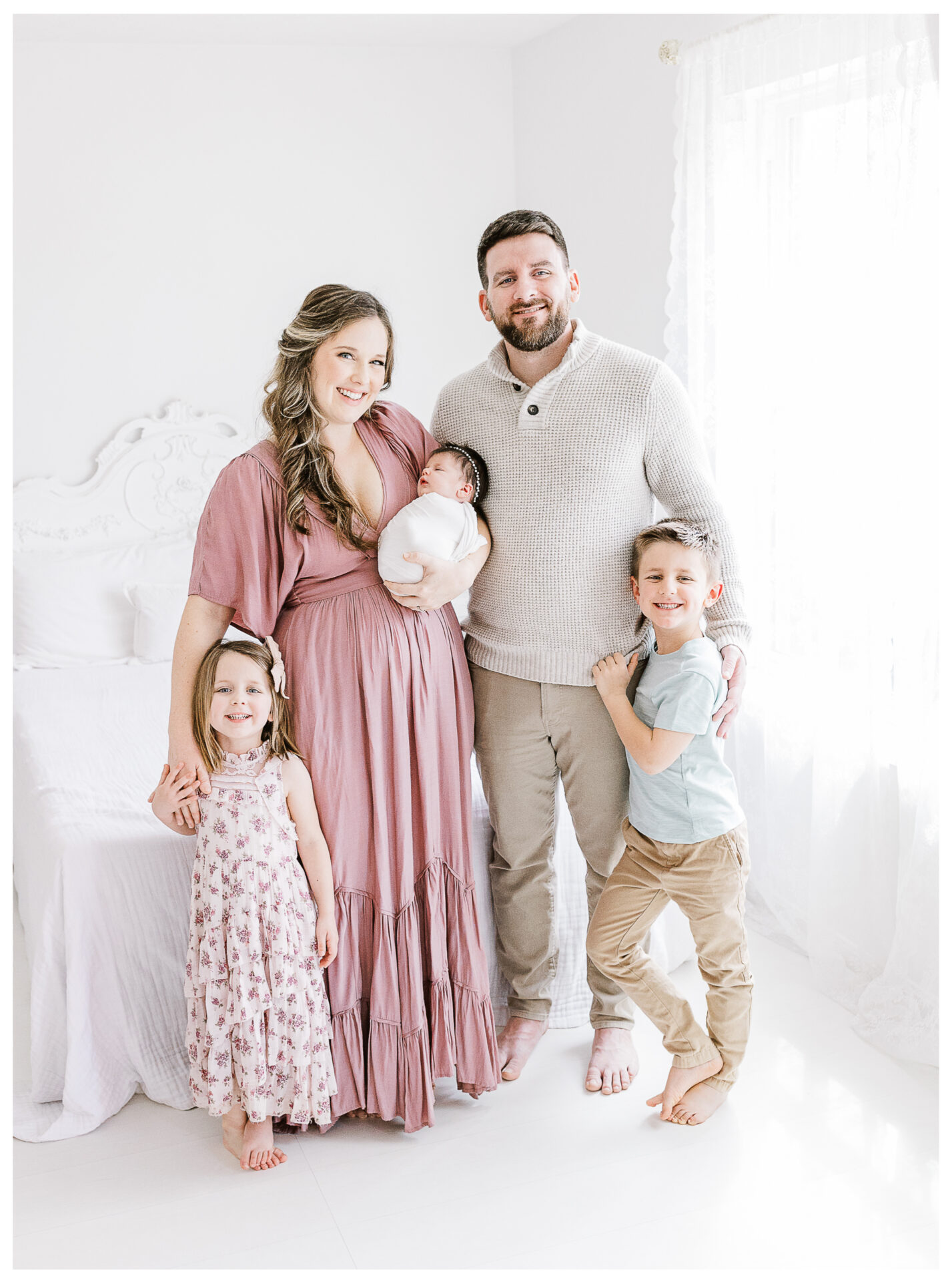 A smiling family of five poses indoors. The mother in a pink dress holds a baby, standing next to the father in a light sweater. Two young children, a girl in a floral dress and a boy in light clothes, stand in front.