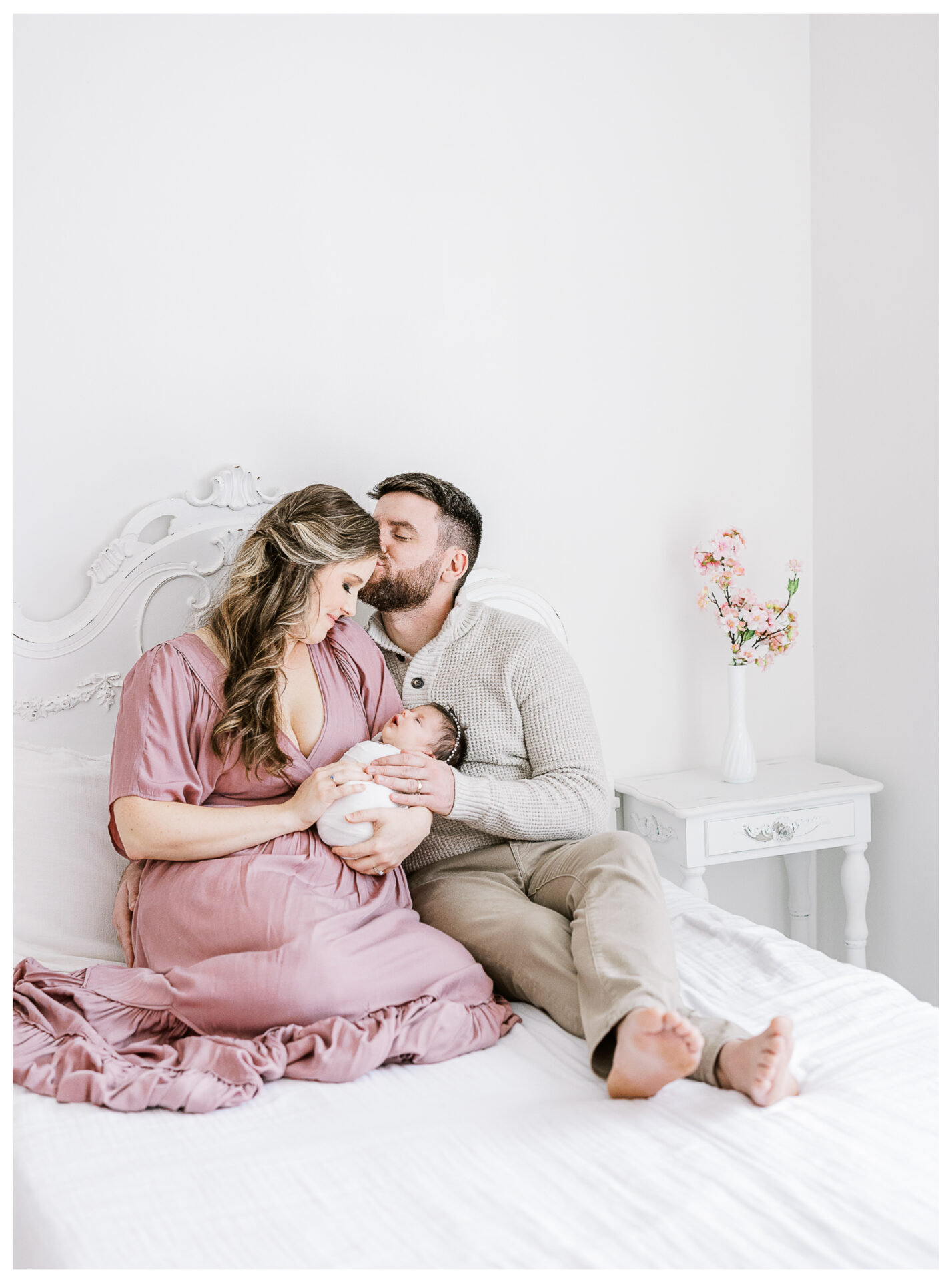 A mother in a mauve dress and a father in a light sweater sit on a white bed, holding and looking lovingly at a newborn baby. The man kisses the woman’s head. A nightstand with a vase of flowers is beside them.