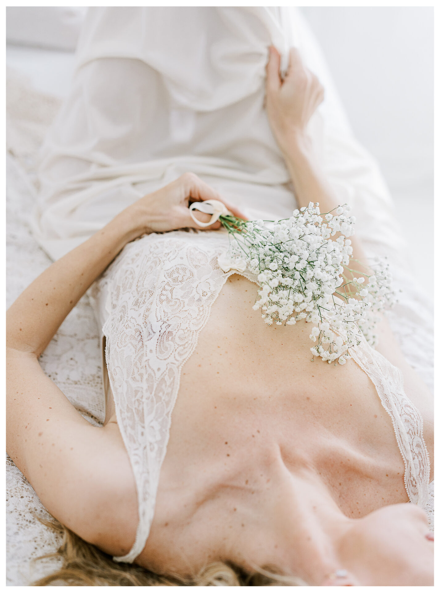 A woman in a delicate lace dress lies on a bed, holding a small bouquet of white baby's breath flowers across her chest, with soft natural light illuminating the scene. Dayton Ohio a romantic blend of timeless beauty and vintage boudoir fashion