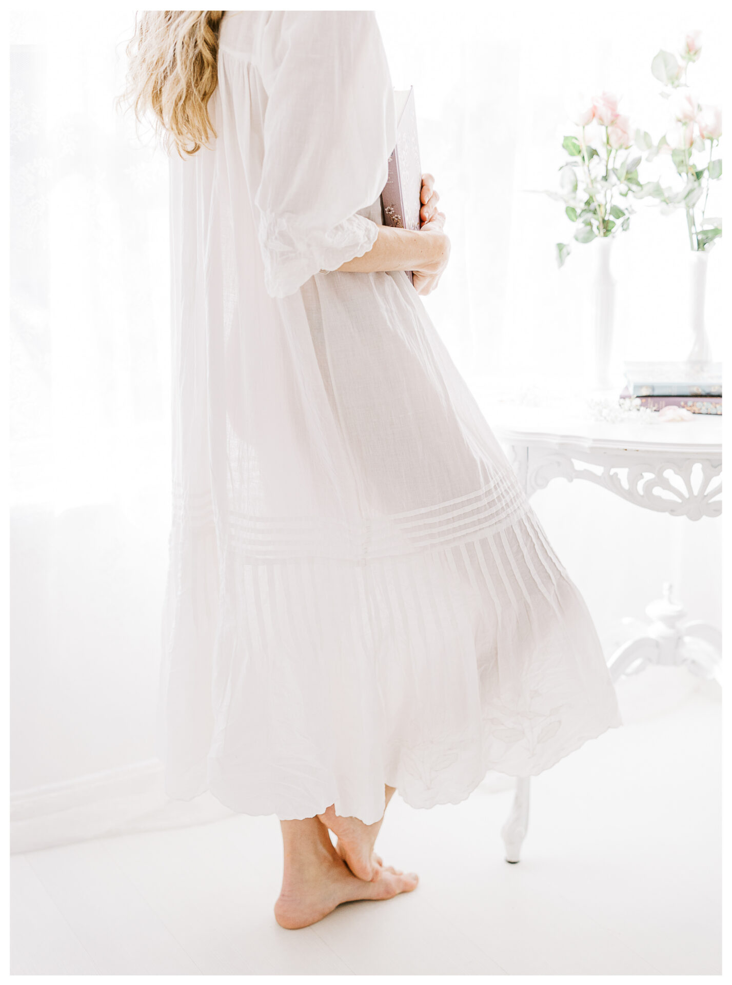 A woman in a long, flowing white dress stands barefoot in a bright, airy room, holding a book. A white table with flowers and books is in the background. Soft natural light fills the scene.