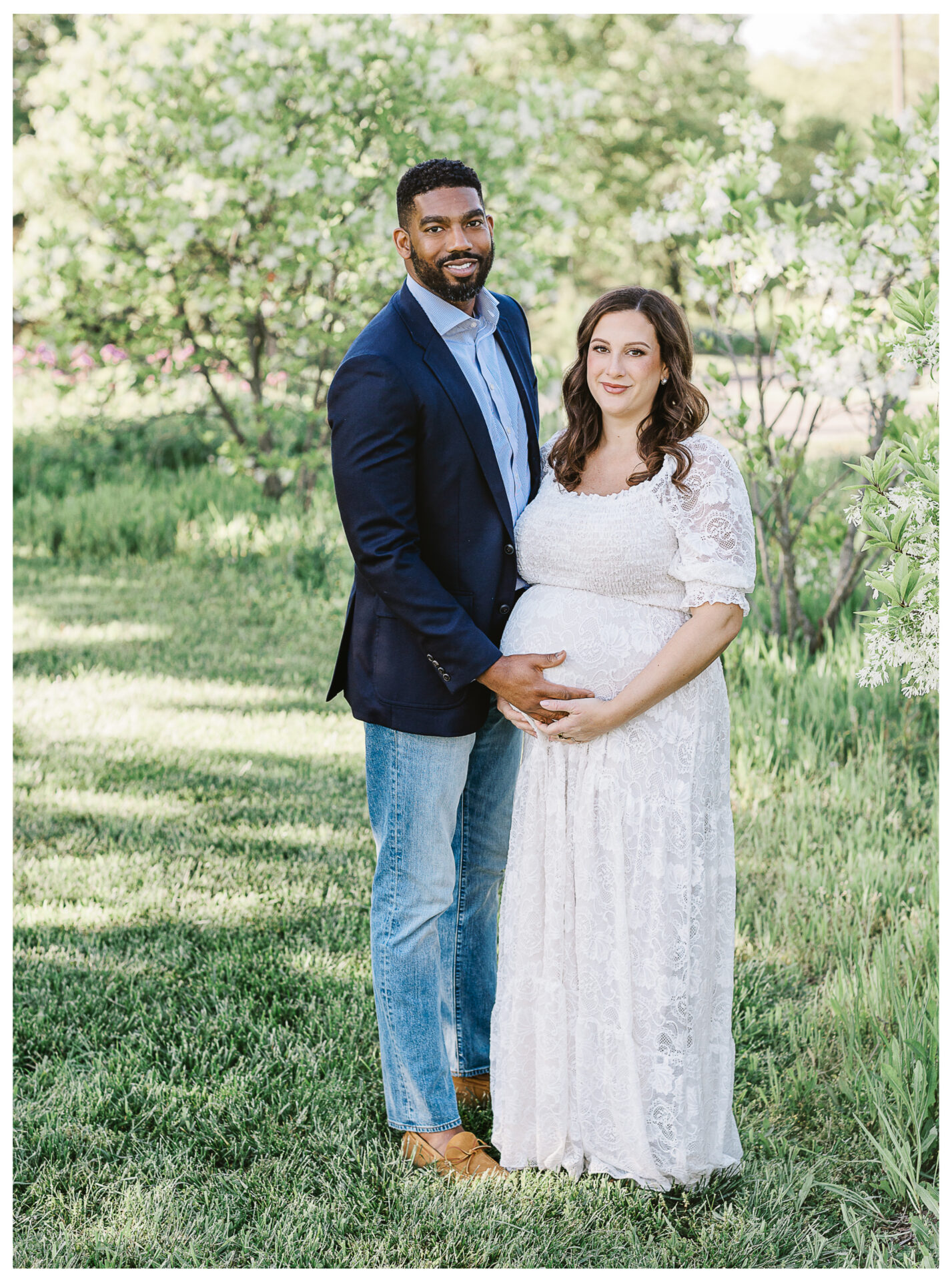 A man in a navy blazer and jeans stands beside a pregnant woman in a white lace dress. They are outdoors in a green, flowered garden, and the man has his hand gently on the woman's baby bump. Both are smiling at the camera.