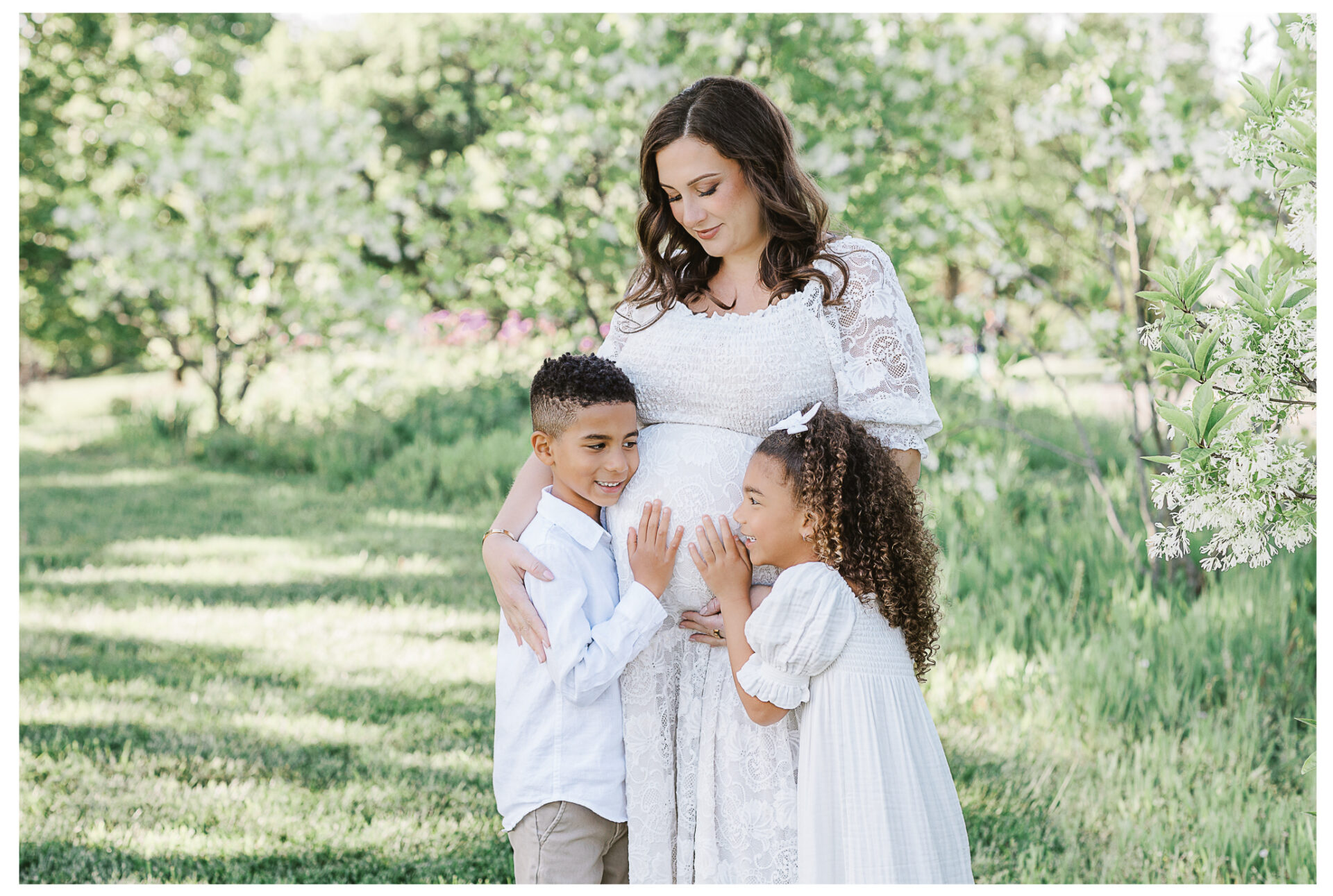 A pregnant mother in a white dress stands in a green, sunlit garden while her young son and daughter, both wearing white, touch her belly and smile, showing affection and excitement.
