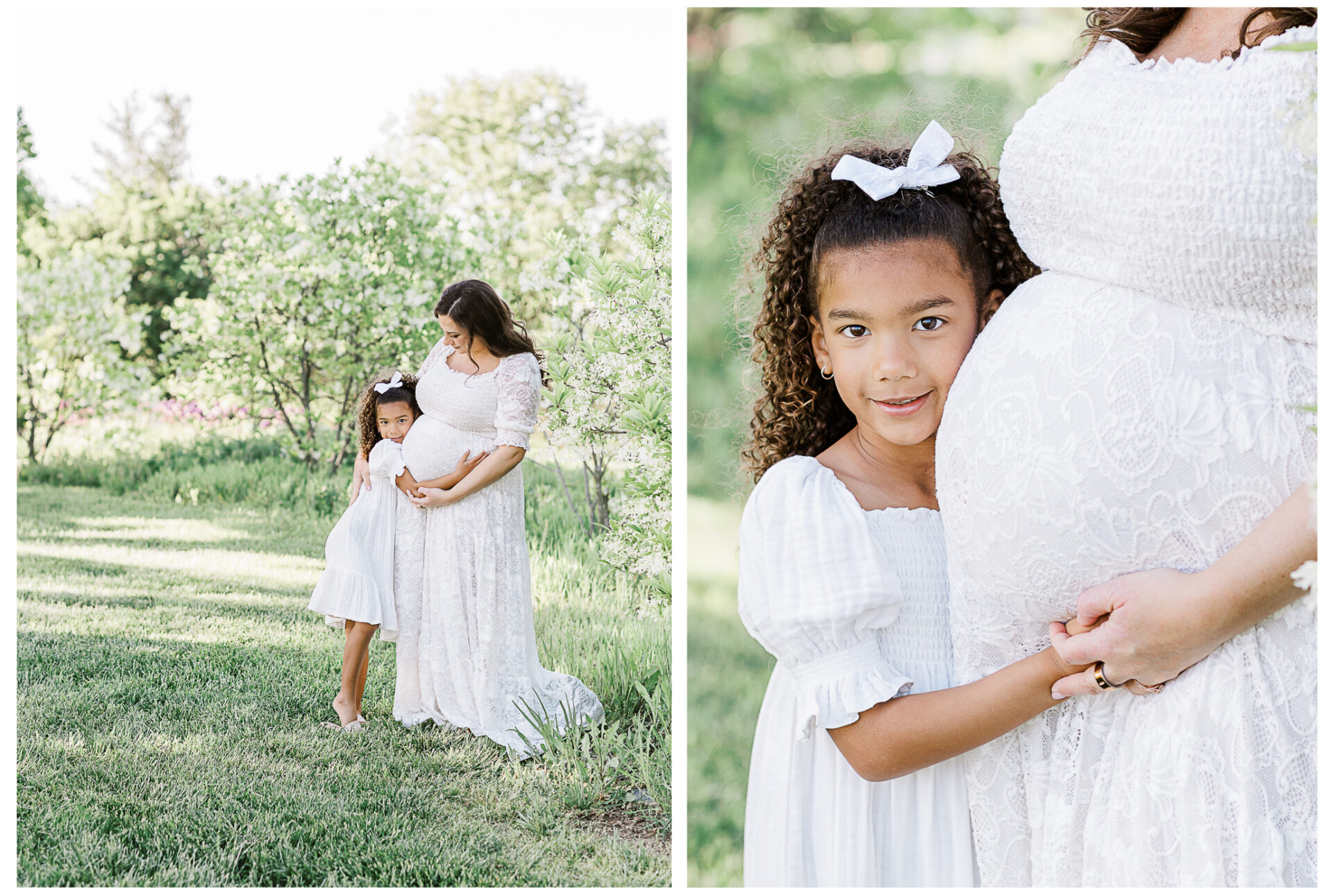A pregnant woman in a white dress stands in a sunlit garden with her young daughter, who gently hugs her baby bump and smiles at the camera. Both wear white dresses and the girl has a white bow in her curly hair.