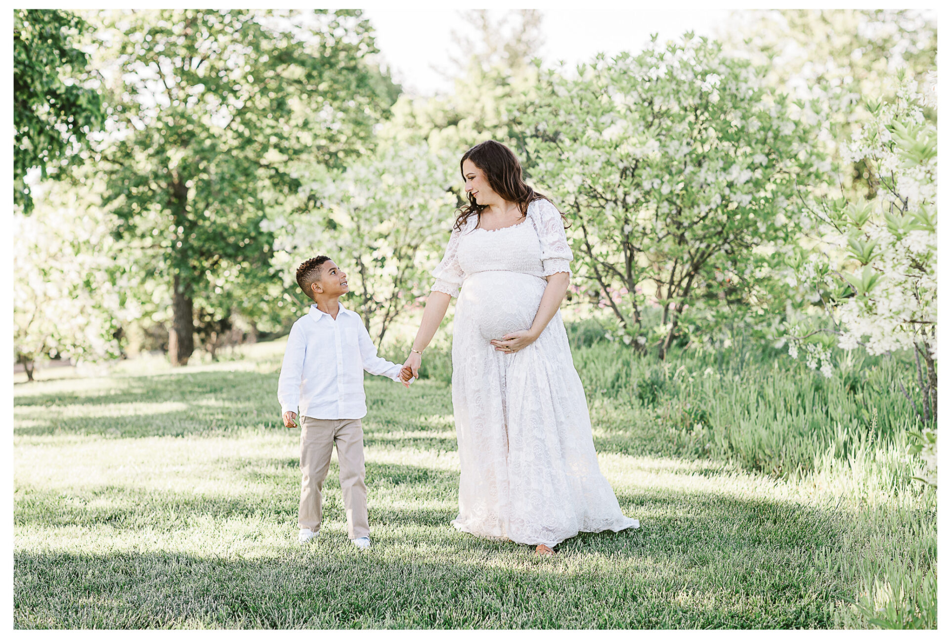 A pregnant mother in a white dress holds hands with her young son in a white shirt and beige pants as they smile at each other, standing on grass in a lush, green outdoor setting.