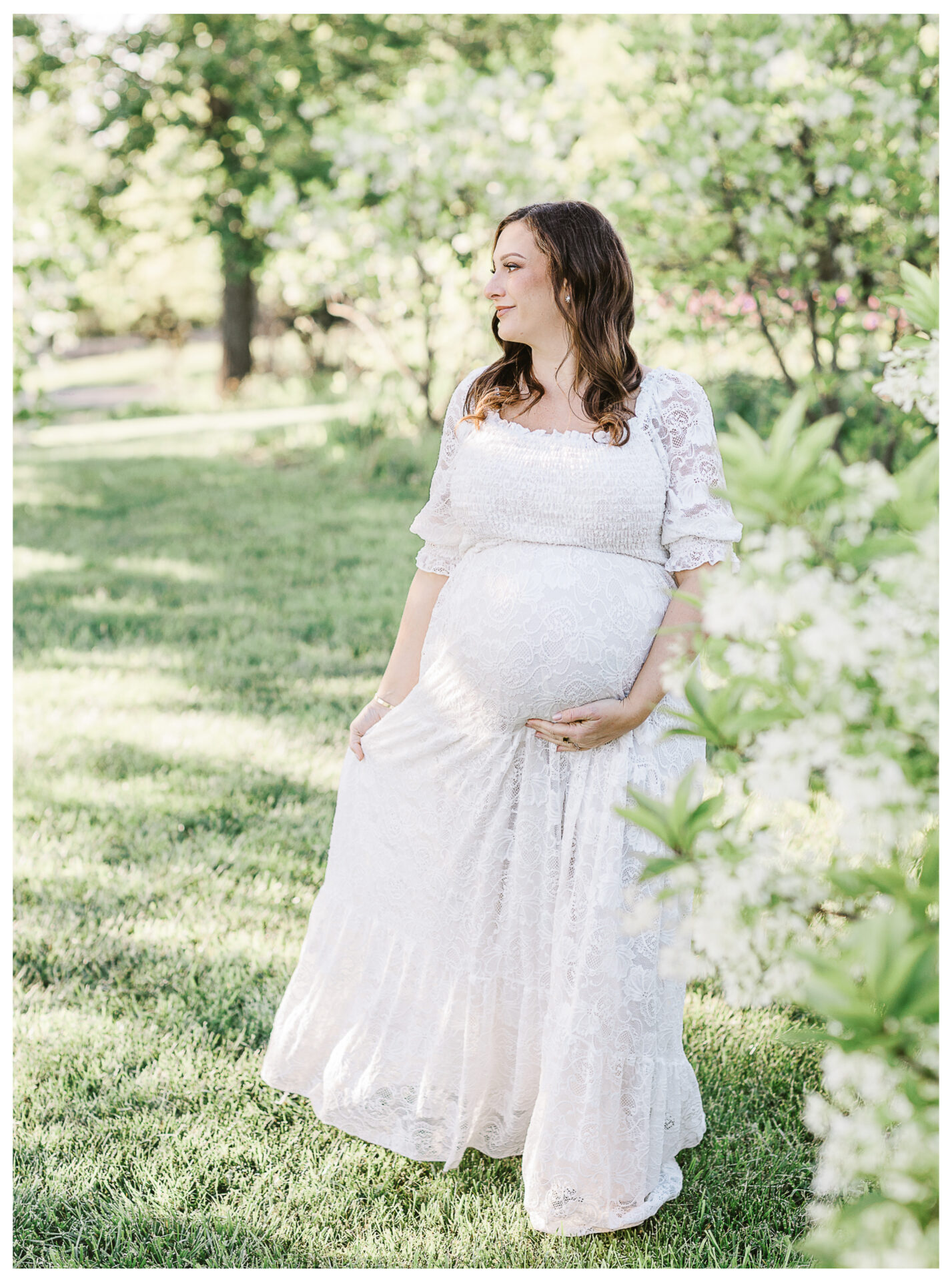 A pregnant woman in a long, white lace dress stands in a sunlit garden, gently holding her belly and looking to the side, surrounded by lush greenery and blooming white flowers.