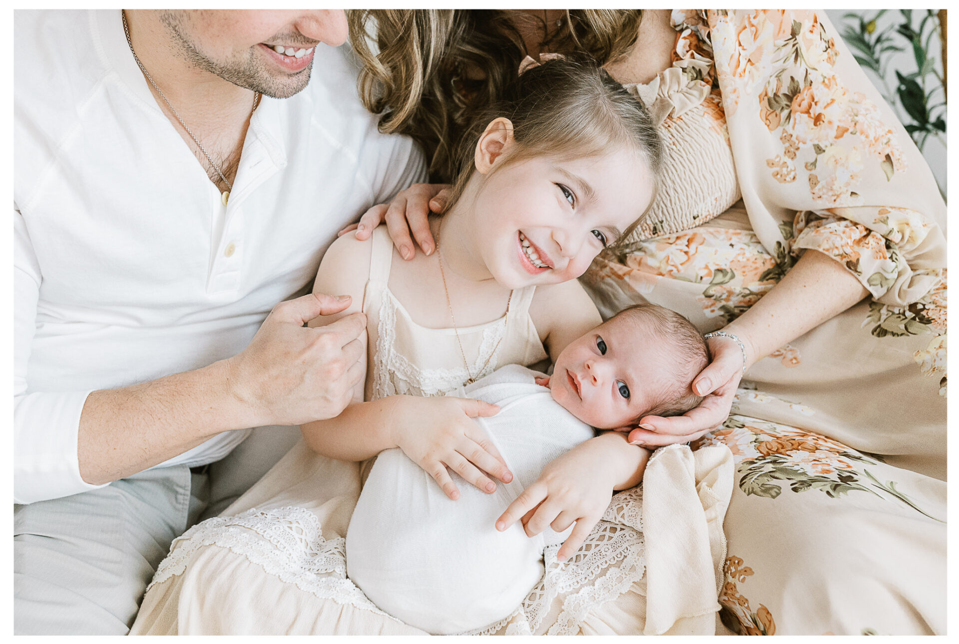 A smiling young girl gently holds her swaddled newborn baby brother while sitting with her parents, who are dressed in light-colored floral and white clothing. The family appears happy and close.