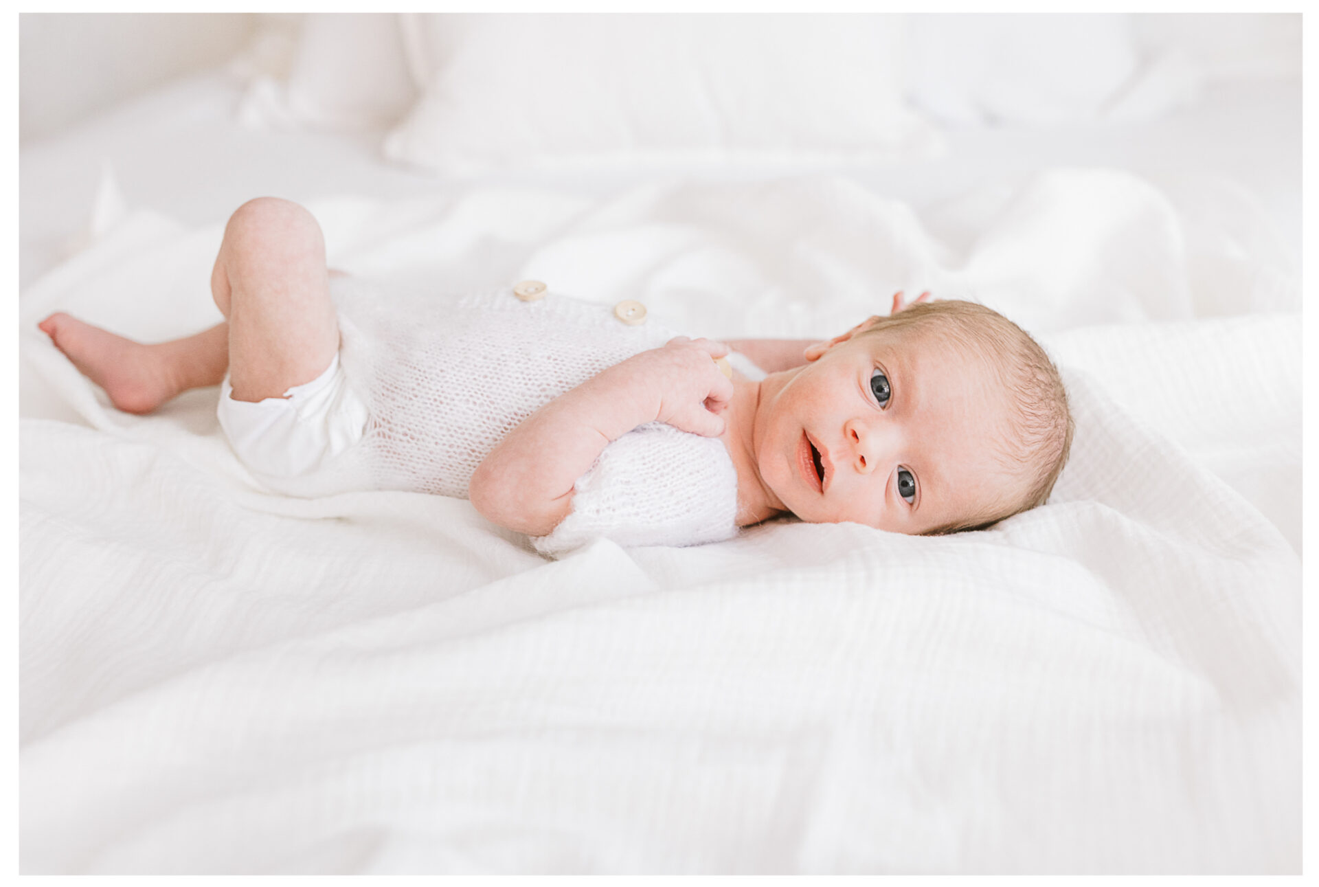 A newborn baby wearing a white outfit lies on a white bed, looking up with wide eyes. The soft bedding and gentle lighting create a calm, peaceful atmosphere.